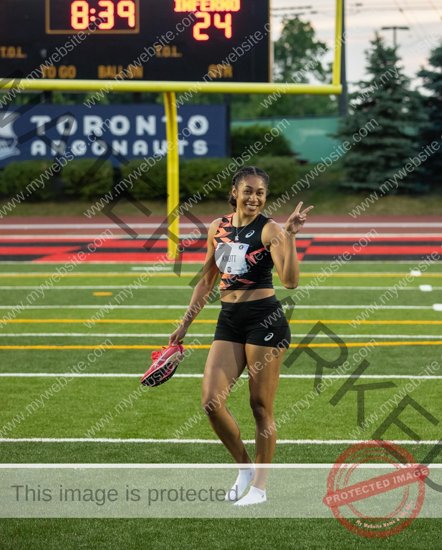 kristina-knott-philippines-knottyourcheese-x-18534 Kristina Knott, track and field star from Philippines, smiles flashing a peace sign by a sports field with her spikes near a scoreboard.