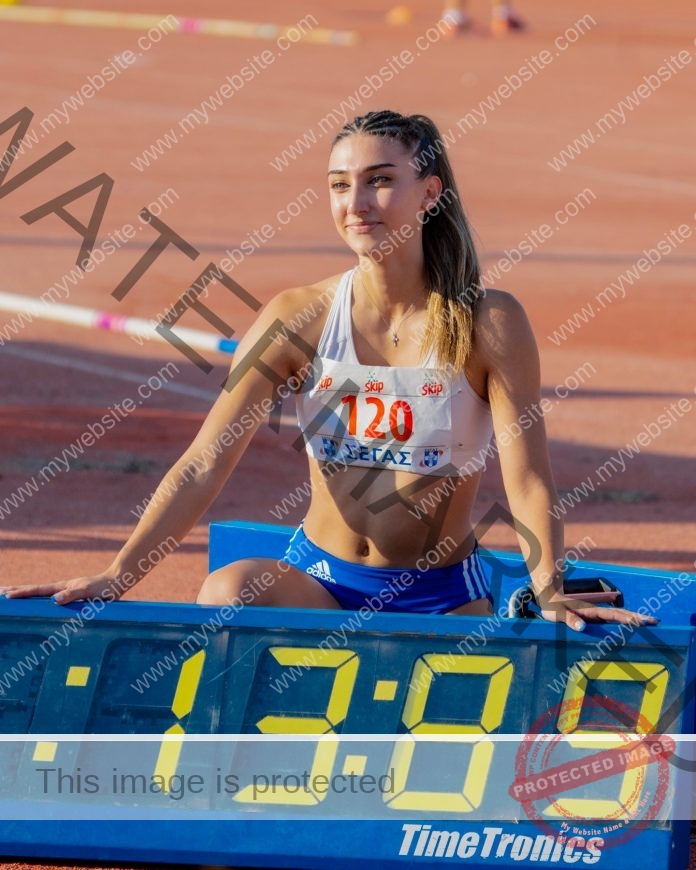 Martha Neamonti, track and field star from Greece, in sportswear and bib 120 smiles kneeling by a timer showing 13:89 on the track.