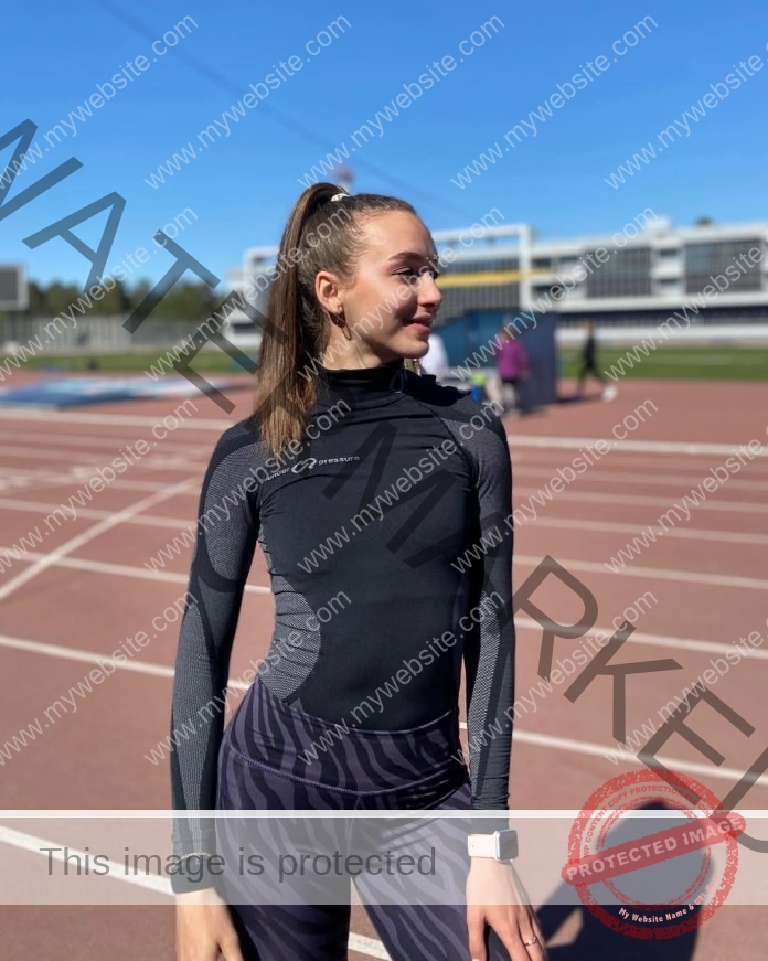 Natalya Kombarova, track and field star from Russia, in athletic wear stands on an outdoor track, smiling with sunlight and sports facilities.
