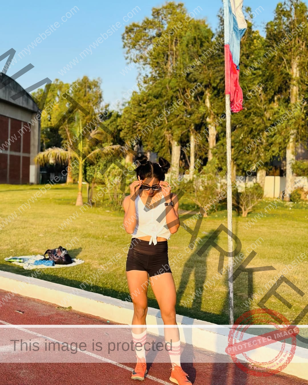 niharika-vashisht-india-niharika.vashhisht-05672 Niharika Vashisht, track and field star from India, stands on a track in sportwear, adjusting sunglasses by a flagpole.