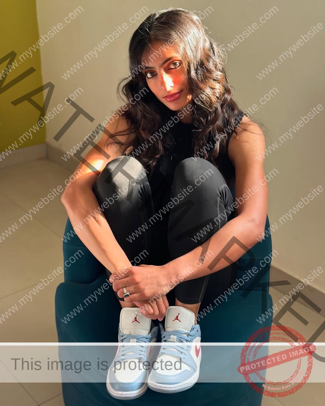 niharika-vashisht-india-niharika.vashhisht-05680 Niharika Vashisht, track and field star from India, with wavy hair sits on a teal chair, hugging her knees in sunlight.
