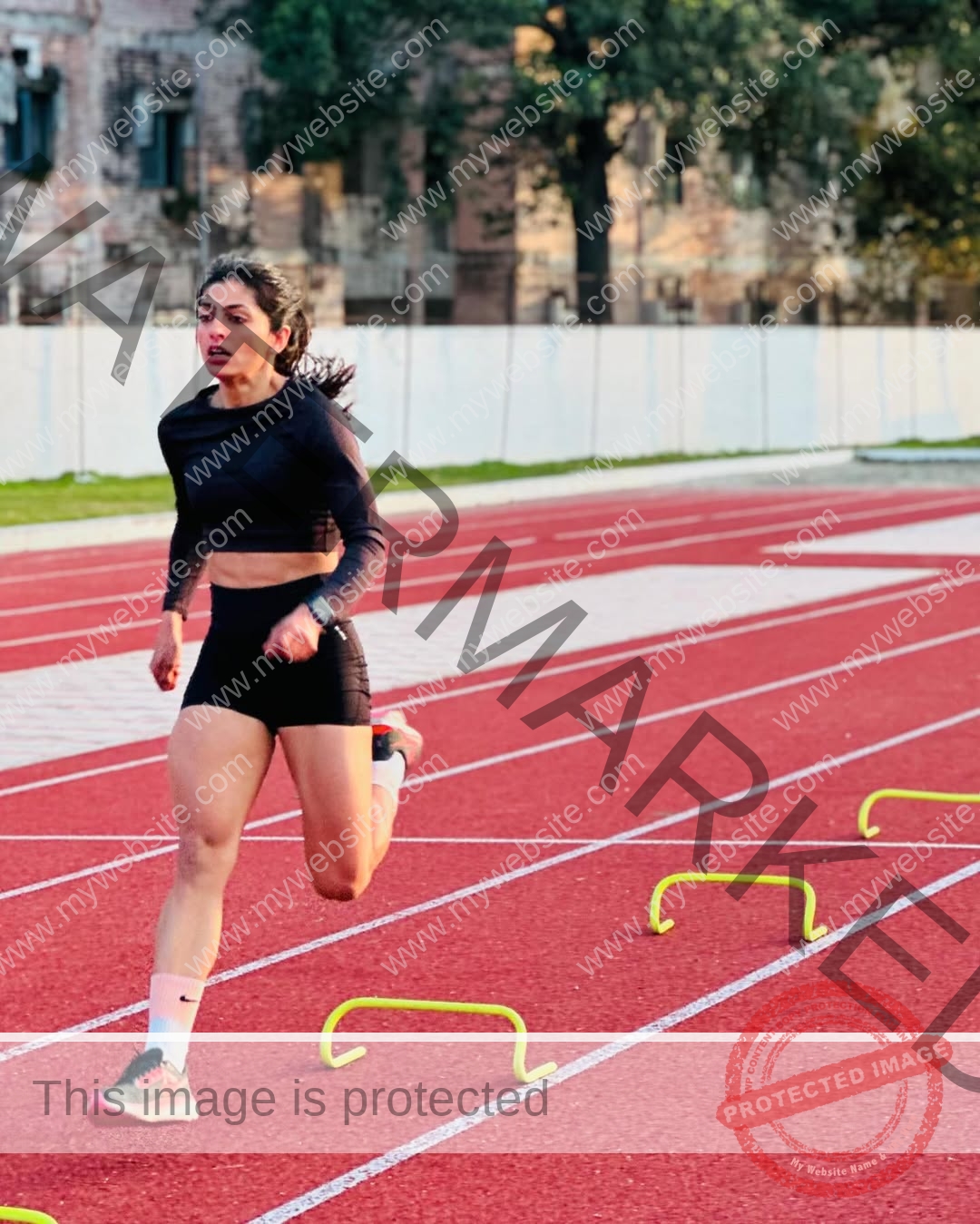 niharika-vashisht-india-niharika.vashhisht-05684 Niharika Vashisht, track and field star from India, runs on a red track leaping over yellow hurdles, with trees and a building behind.