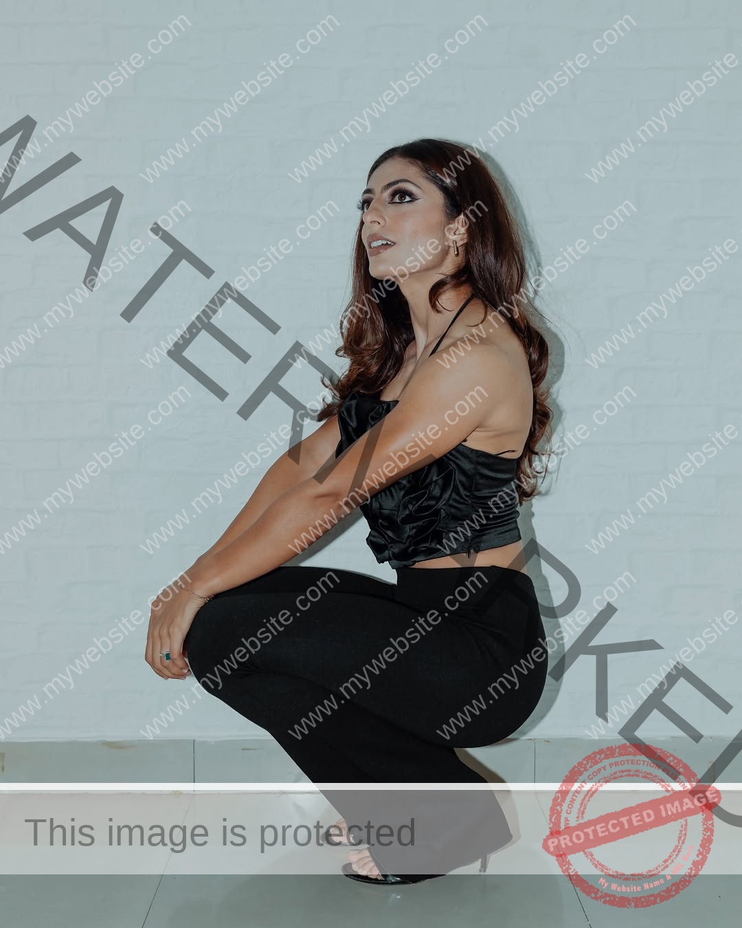 niharika-vashisht-india-niharika.vashhisht-05739 Niharika Vashisht, track and field star from India, with long brown hair in black attire, squats indoors by a white brick wall.