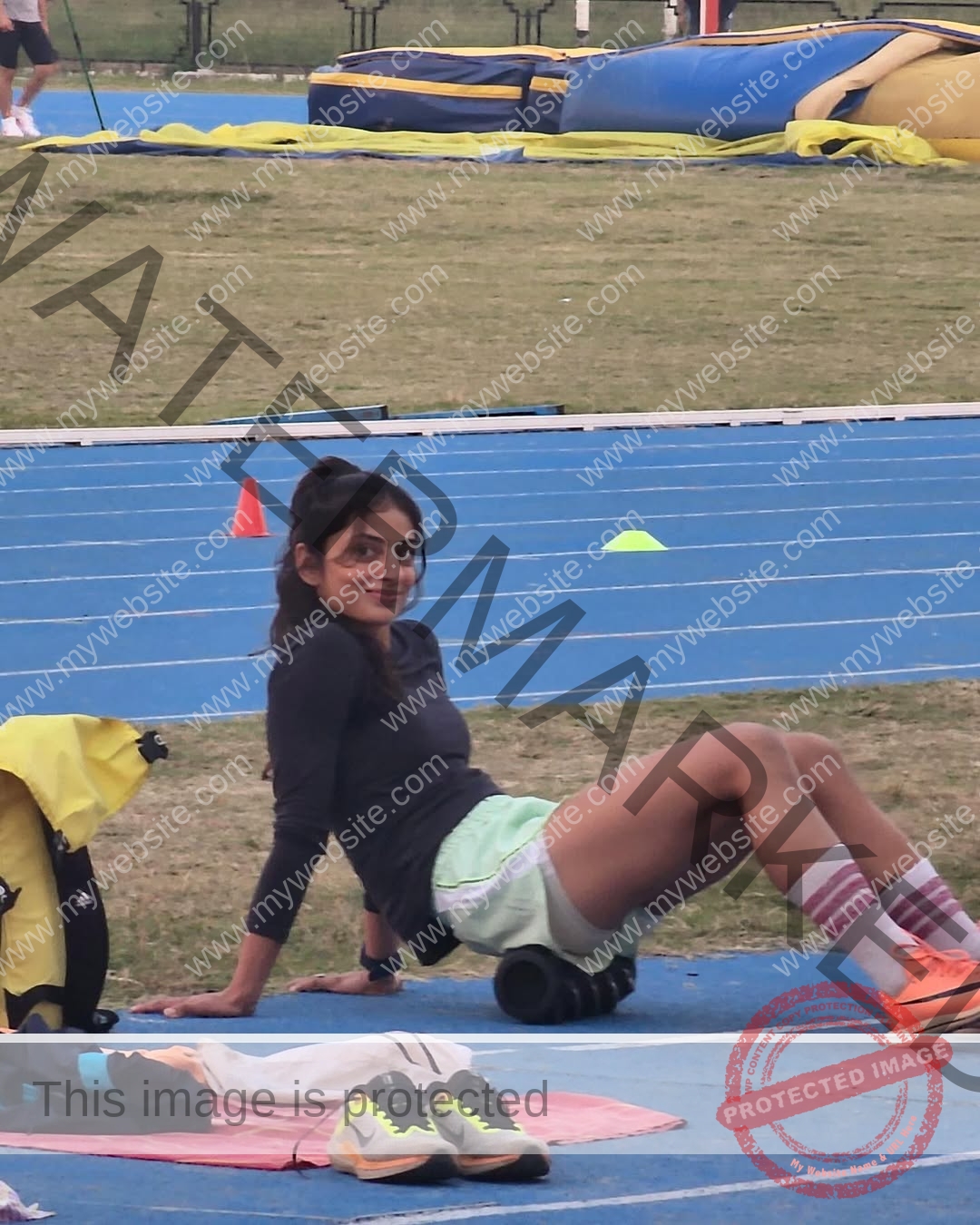niharika-vashisht-india-niharika.vashhisht-05794 Niharika Vashisht, track and field star from India, smiles in athletic wear using a foam roller on a blue track, sports gear nearby.