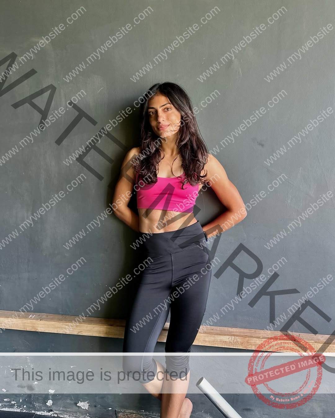 niharika-vashisht-india-niharika.vashhisht-05948 Niharika Vashisht, track and field star from India, with long dark hair in a pink sports bra and black leggings, stands barefoot, arms behind her back, against a gray wall.