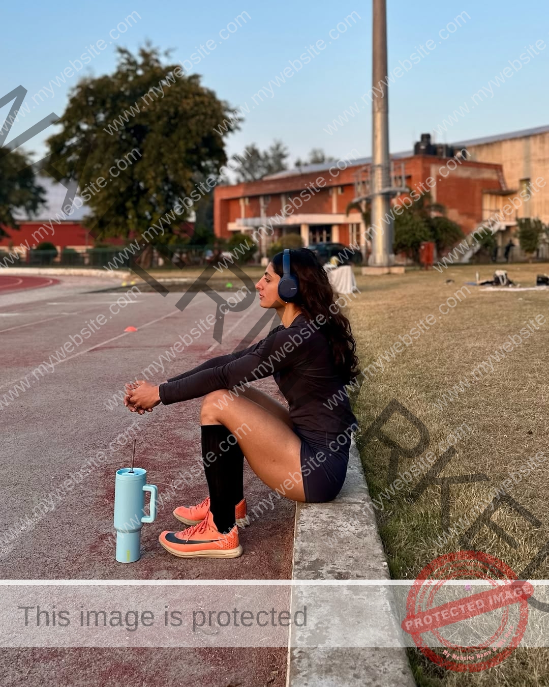 niharika-vashisht-india-niharika.vashhisht-05986 Niharika Vashisht, track and field star from India, in athletic wear and headphones, rests on a curb by a track holding water.