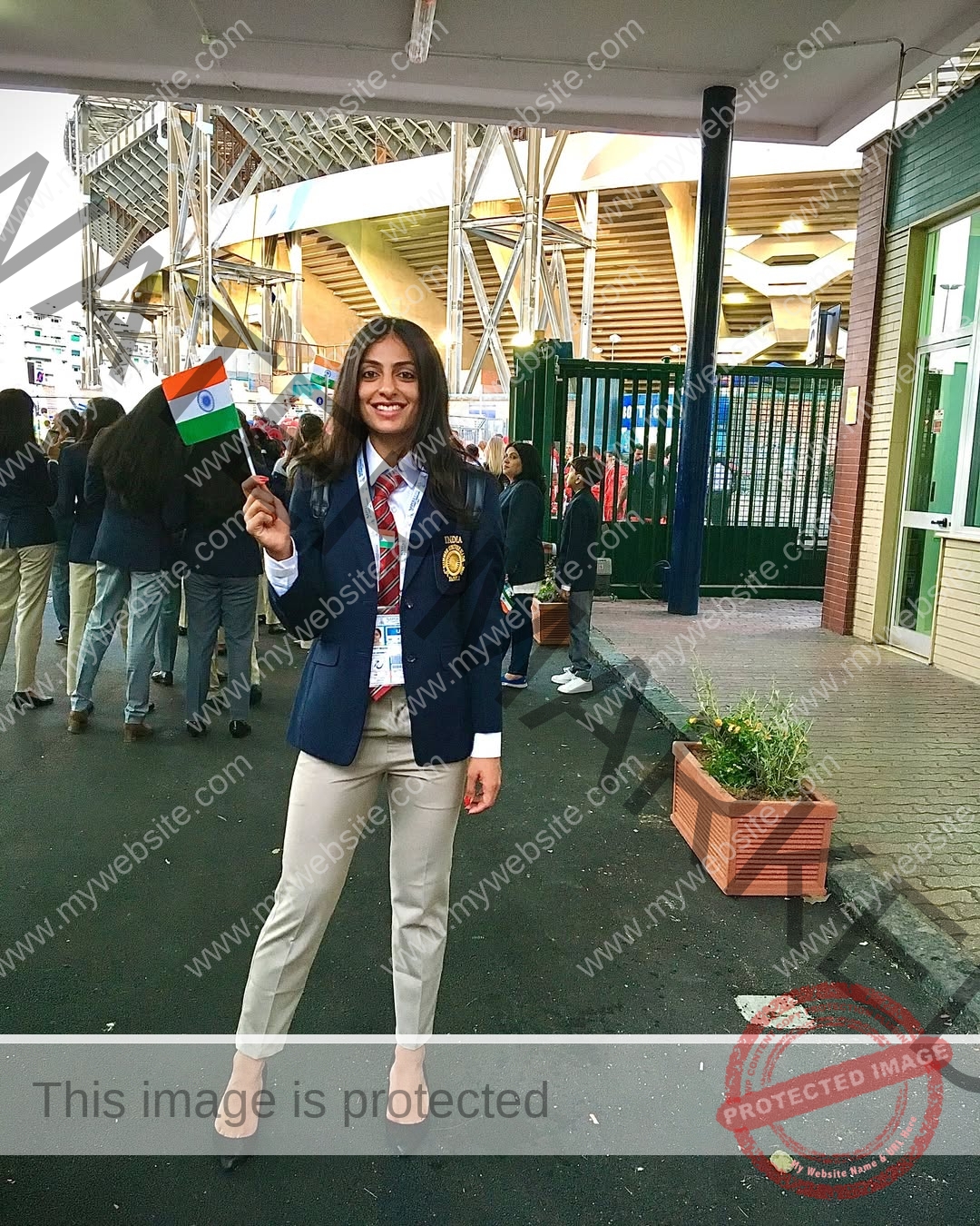 niharika-vashisht-india-niharika.vashhisht-05990 Niharika Vashisht, track and field star from India, smiles in a formal blazer, holding the Indian flag near a stadium with others.