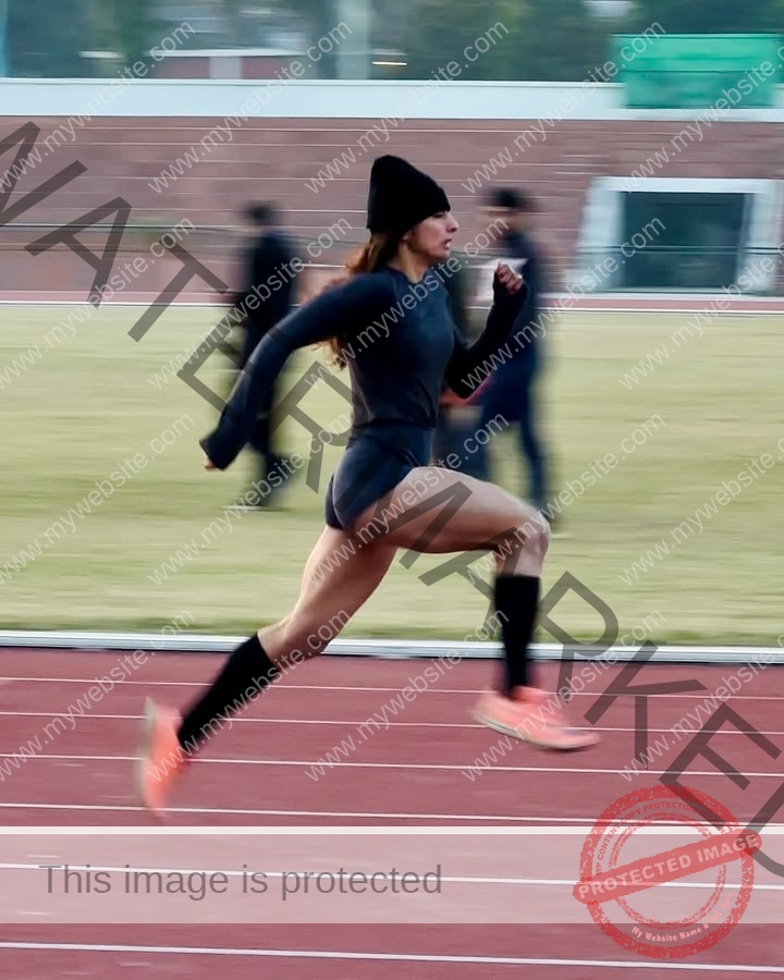 niharika-vashisht-india-niharika.vashhisht-05995 Niharika Vashisht, track and field star from India sprints in black outfit and beanie on a track, mid-stride, blurred field behind.