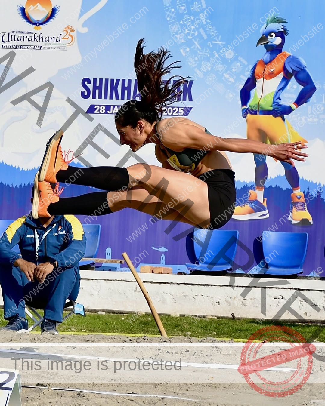 niharika-vashisht-india-niharika.vashhisht-06003 Niharika Vashisht, track and field star from India leaps mid-air in long jump as blue mascot, banner, sandpit & officials appear nearby.