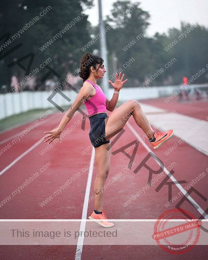 niharika-vashisht-india-niharika.vashhisht-06032 Niharika Vashisht, track and field star from India, does high knees on an outdoor track with trees and others in the background.