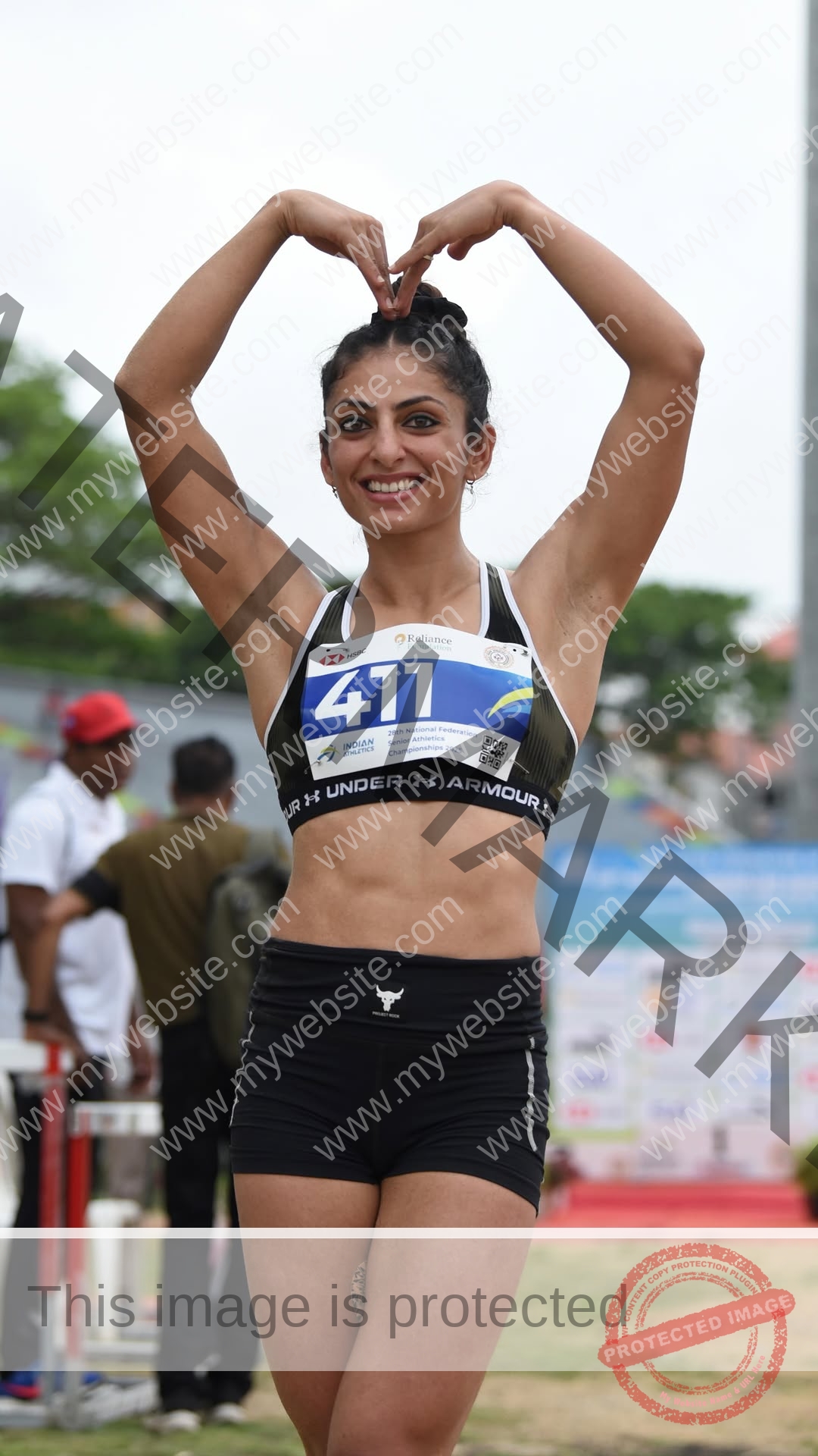 niharika-vashisht-india-niharika.vashhisht-06077 Niharika Vashisht, track and field star from India stands outdoors in sportswear, smiling, arms above head in heart shape, race bib 411.