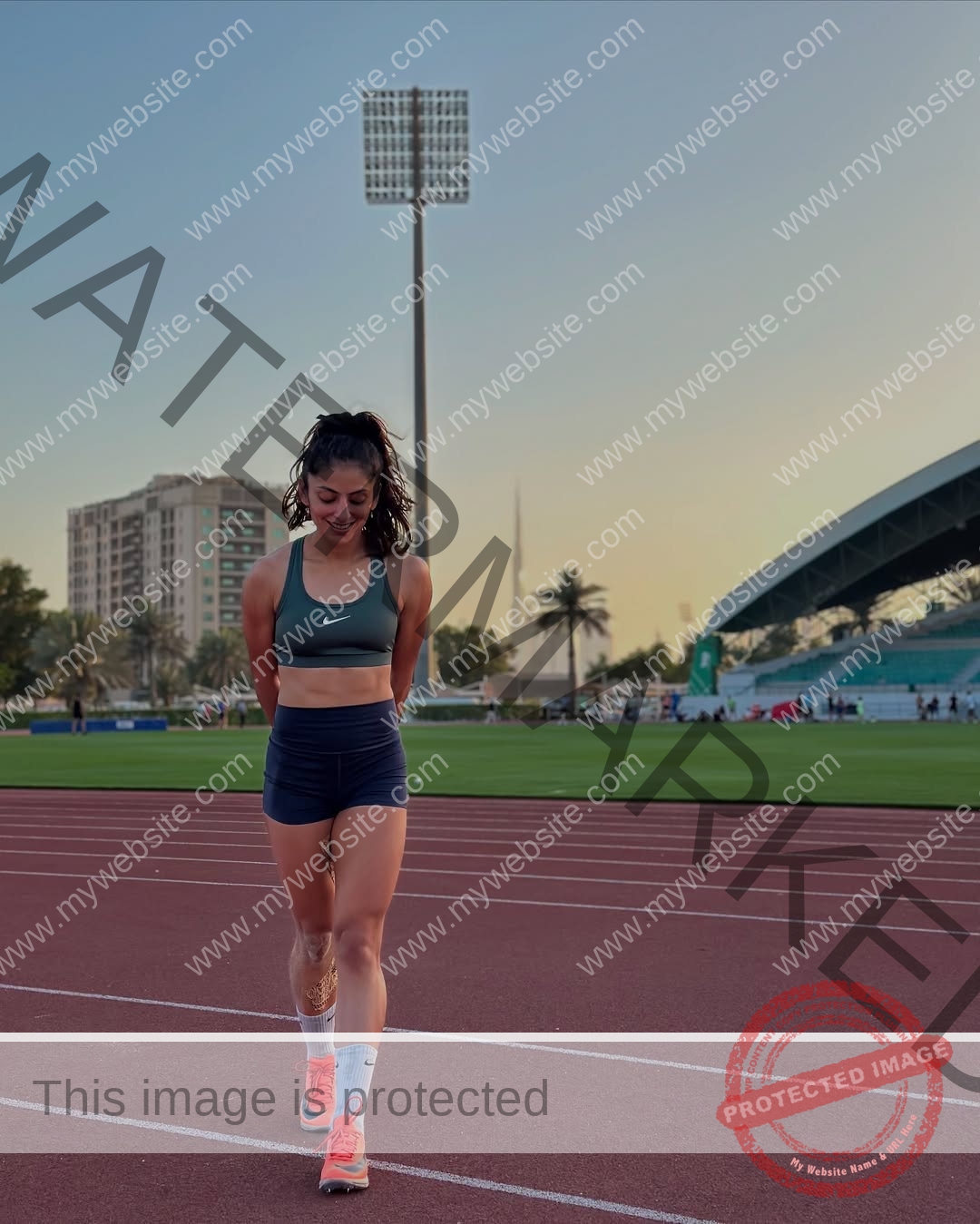 niharika-vashisht-india-niharika.vashhisht-06089 Niharika Vashisht, track and field star from India, walks on a running track at sunset in sportswear; stadium and palm trees behind.