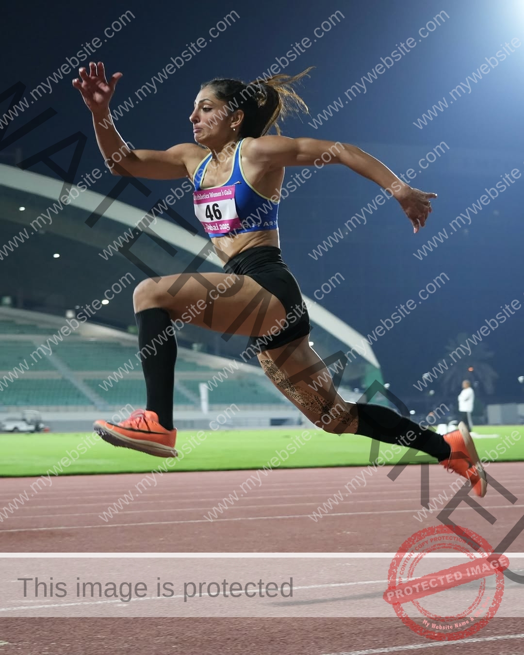 niharika-vashisht-india-niharika.vashhisht-06096 Niharika Vashisht, track and field star from India, in mid-air during a long jump at night, blue sports bra, black shorts, stadium lights.
