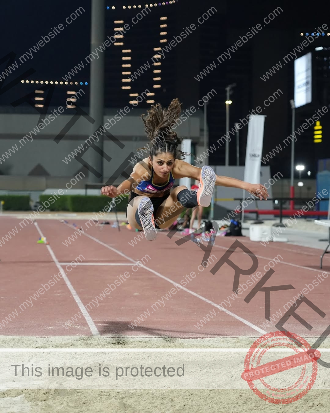 niharika-vashisht-india-niharika.vashhisht-06097 Niharika Vashisht, track and field star from India, is mid-air performing a long jump at night with sand and city lights behind her.