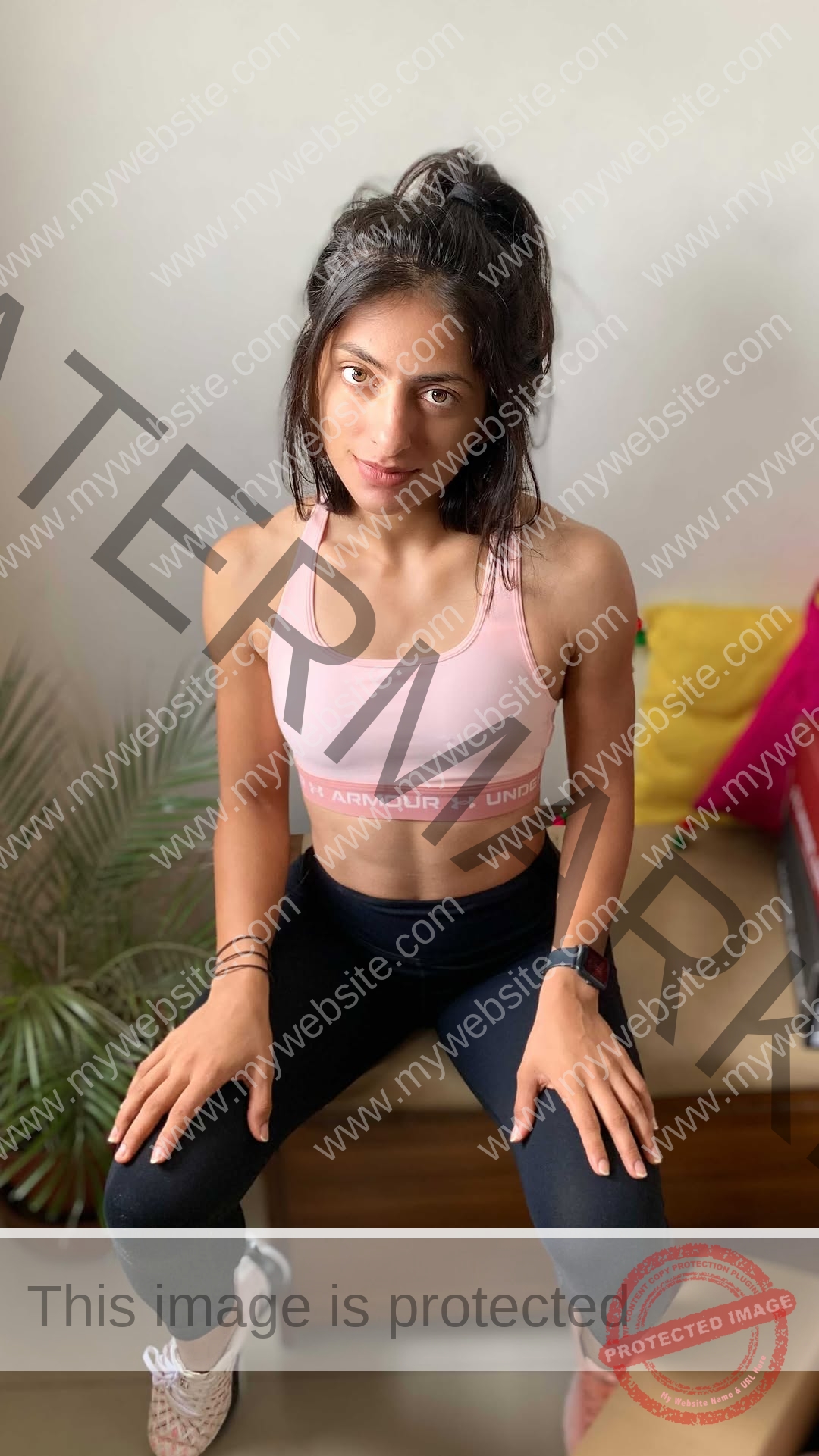 niharika-vashisht-india-niharika.vashhisht-06101 Niharika Vashisht, track and field star from India, with dark hair in a ponytail, wears a pink sports bra and black leggings on a bench indoors; green plant and colorful pillows in background.
