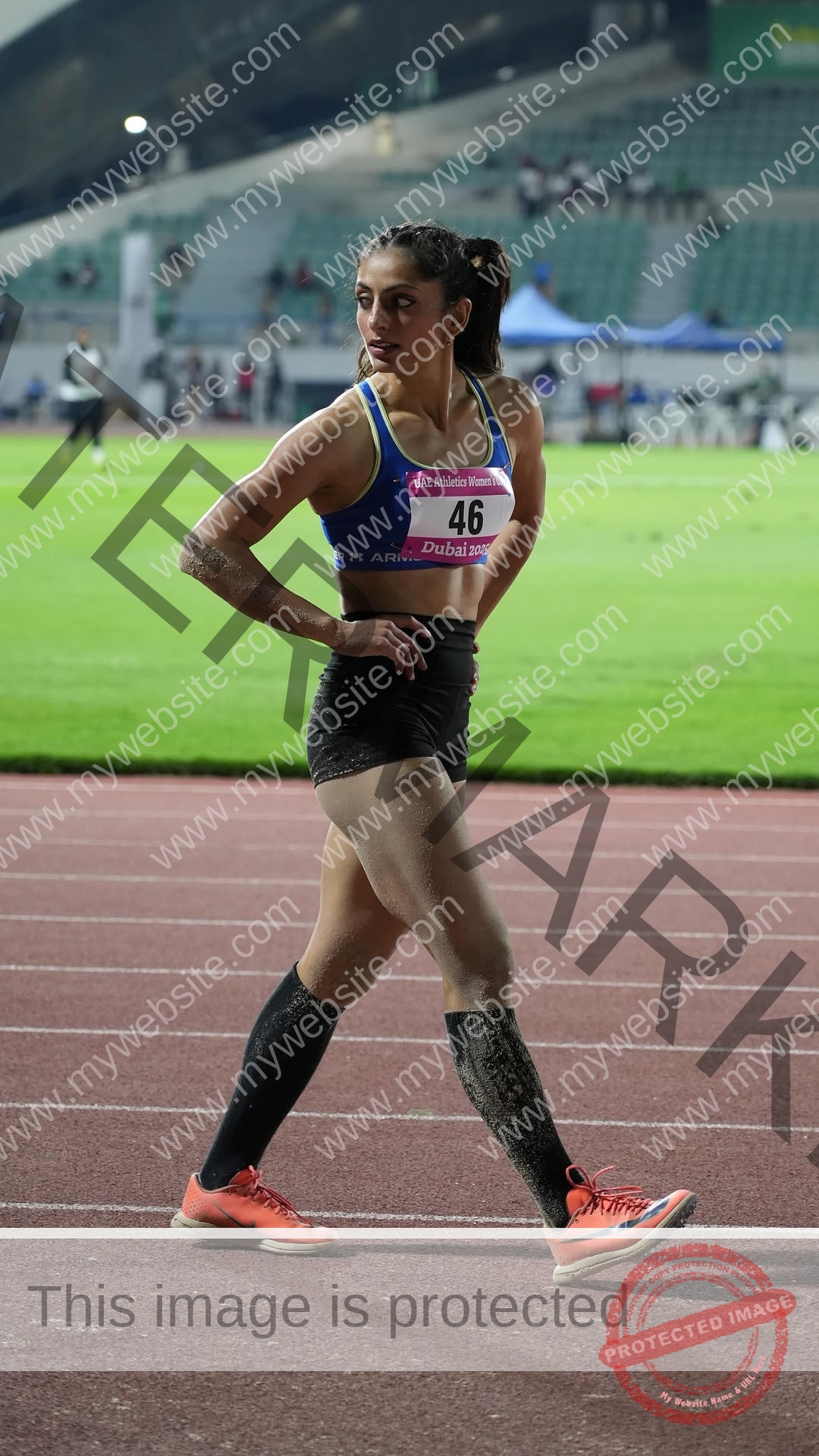 niharika-vashisht-india-niharika.vashhisht-06108 Niharika Vashisht, track and field star from India, stands on the track, number 46 bib, sandy from a jump. Lit stadium, fans behind.