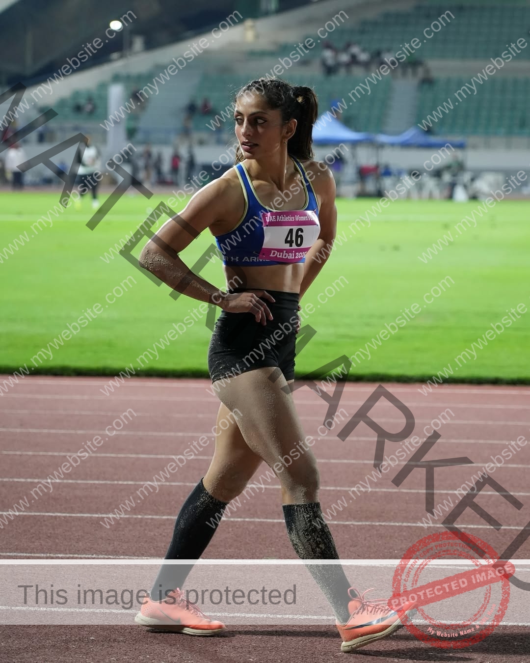 niharika-vashisht-india-niharika.vashhisht-06112 Niharika Vashisht, track and field star from India, stands on the track in blue sports bra and black shorts, sandy legs, looking aside.