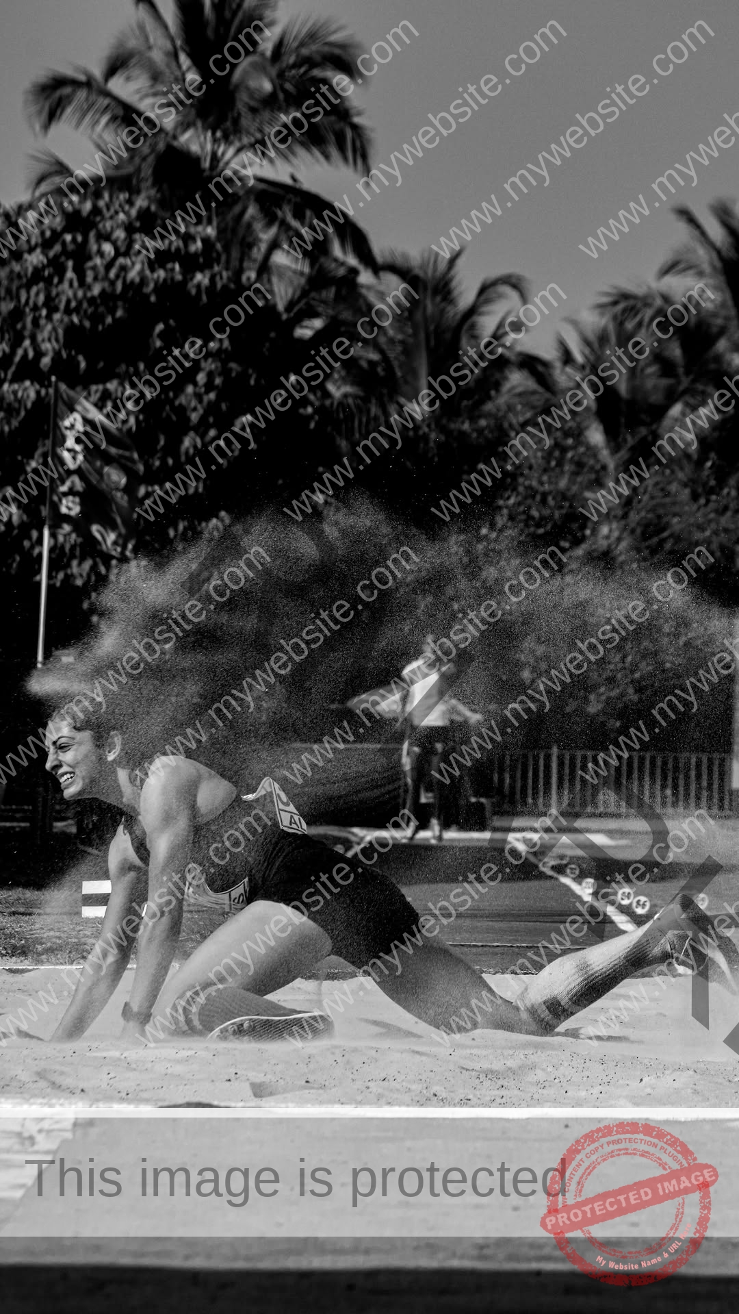 niharika-vashisht-india-niharika.vashhisht-06133 Niharika Vashisht, track and field star from India lands in sand during a long jump; sand flies up, palm trees and a person visible.