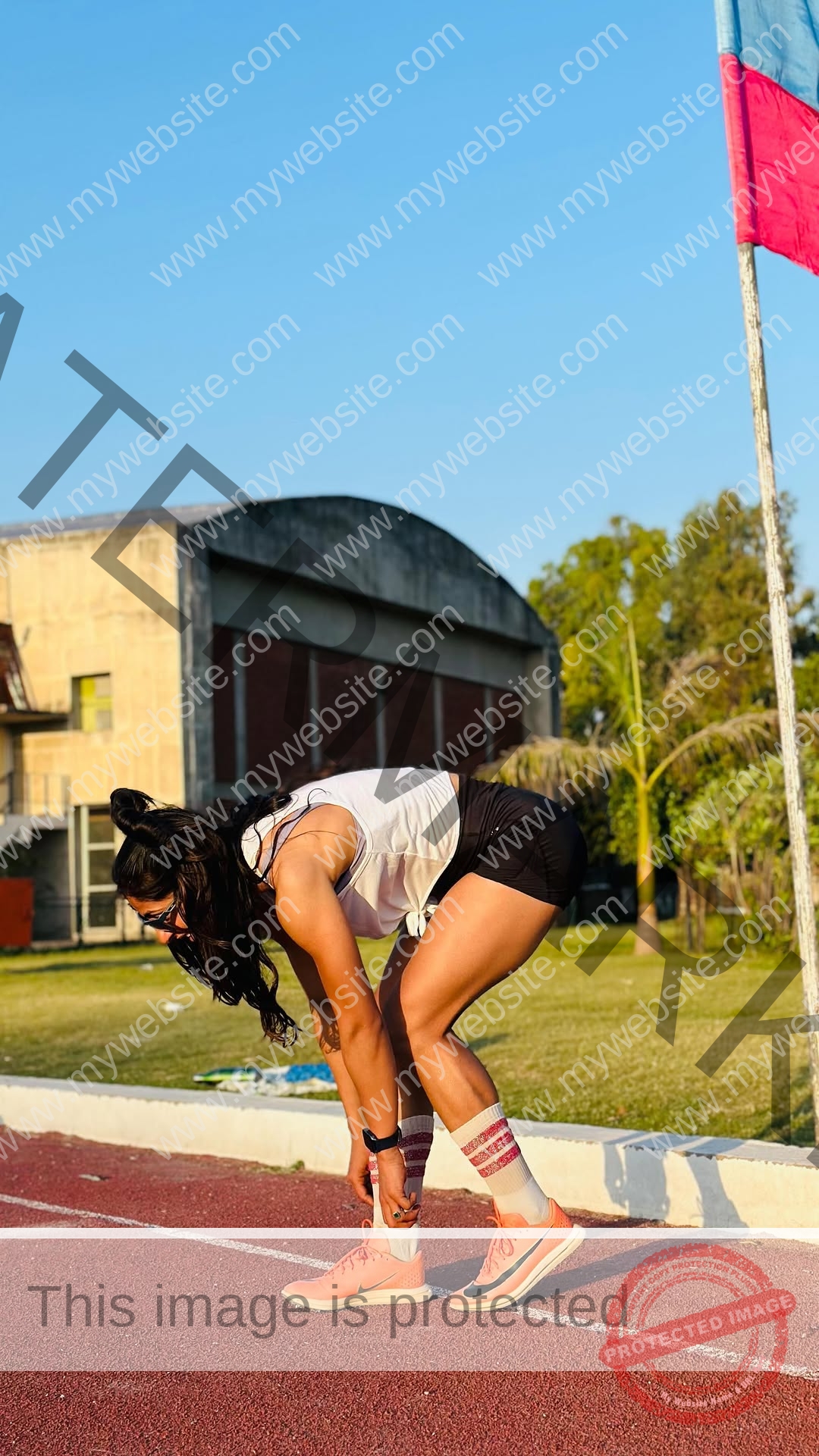 niharika-vashisht-india-niharika.vashhisht-06179 Niharika Vashisht, track and field star from India bends over on a track to stretch or catch her breath near grass, trees, and a building.