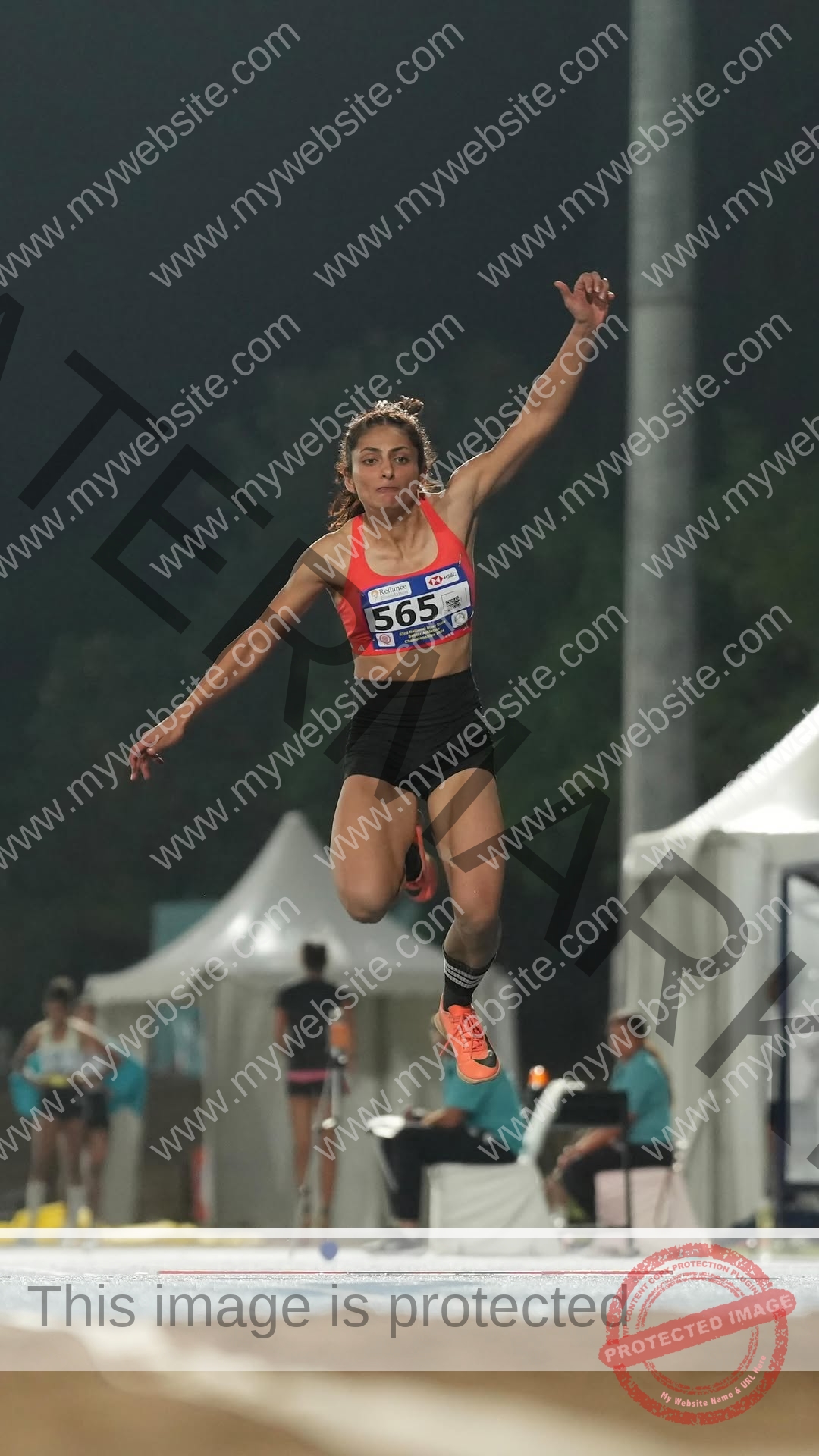 niharika-vashisht-india-niharika.vashhisht-06184 Niharika Vashisht, track and field star from India leaps in mid-air wearing a red sports bra, black shorts, race bib 565 at night.