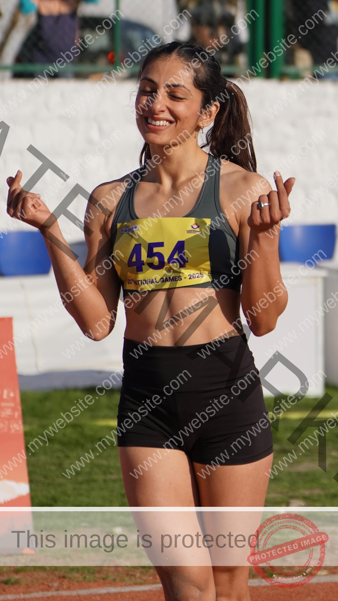 niharika-vashisht-india-niharika.vashhisht-06220 Niharika Vashisht, track and field star from India, smiles on a track in bib 454, eyes closed, making finger hearts with both hands.