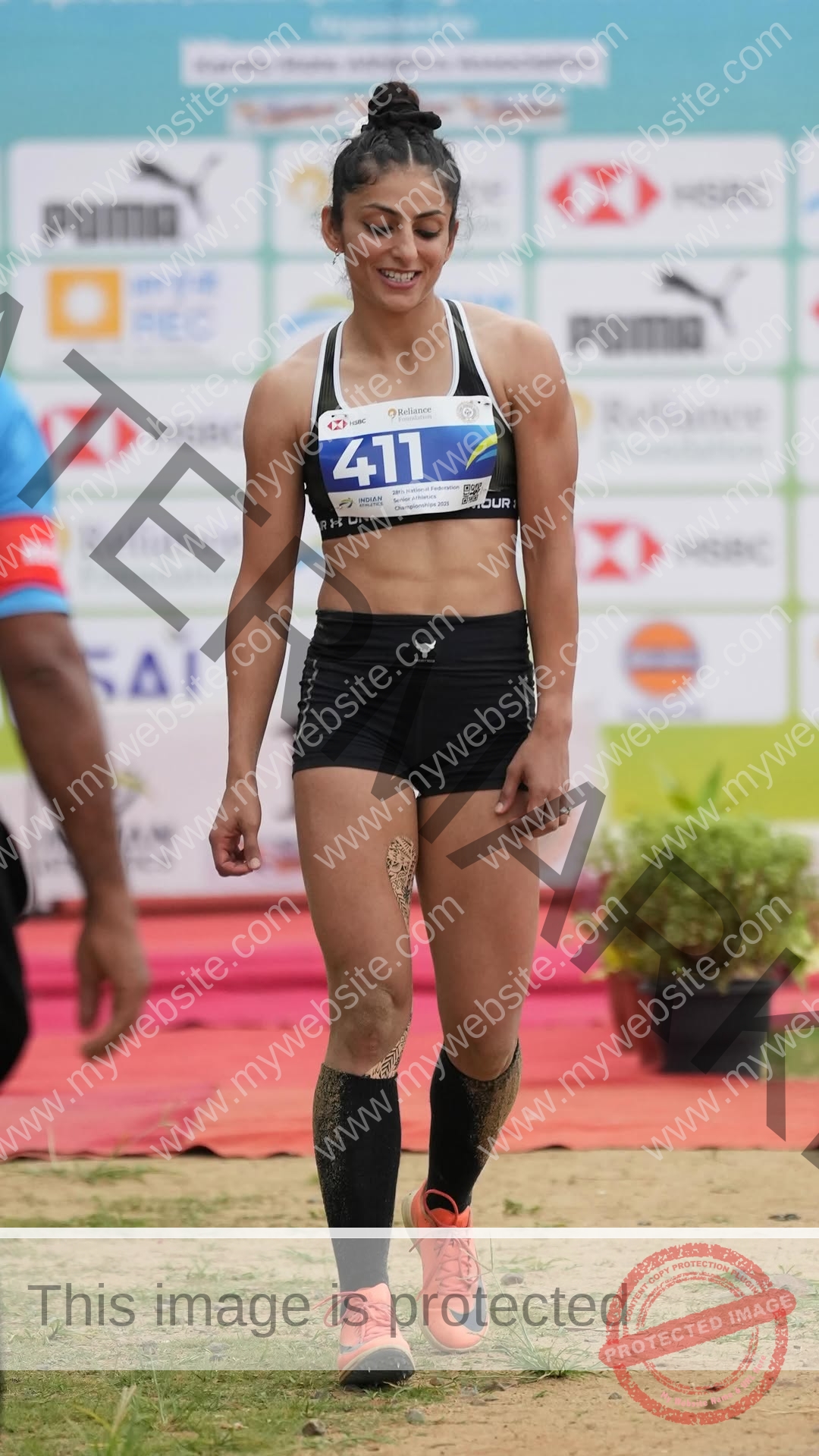 niharika-vashisht-india-niharika.vashhisht-06221 Niharika Vashisht, track and field star from India, wearing black sportswear and orange shoes, smiles walking on track; bib 411, thigh taped.