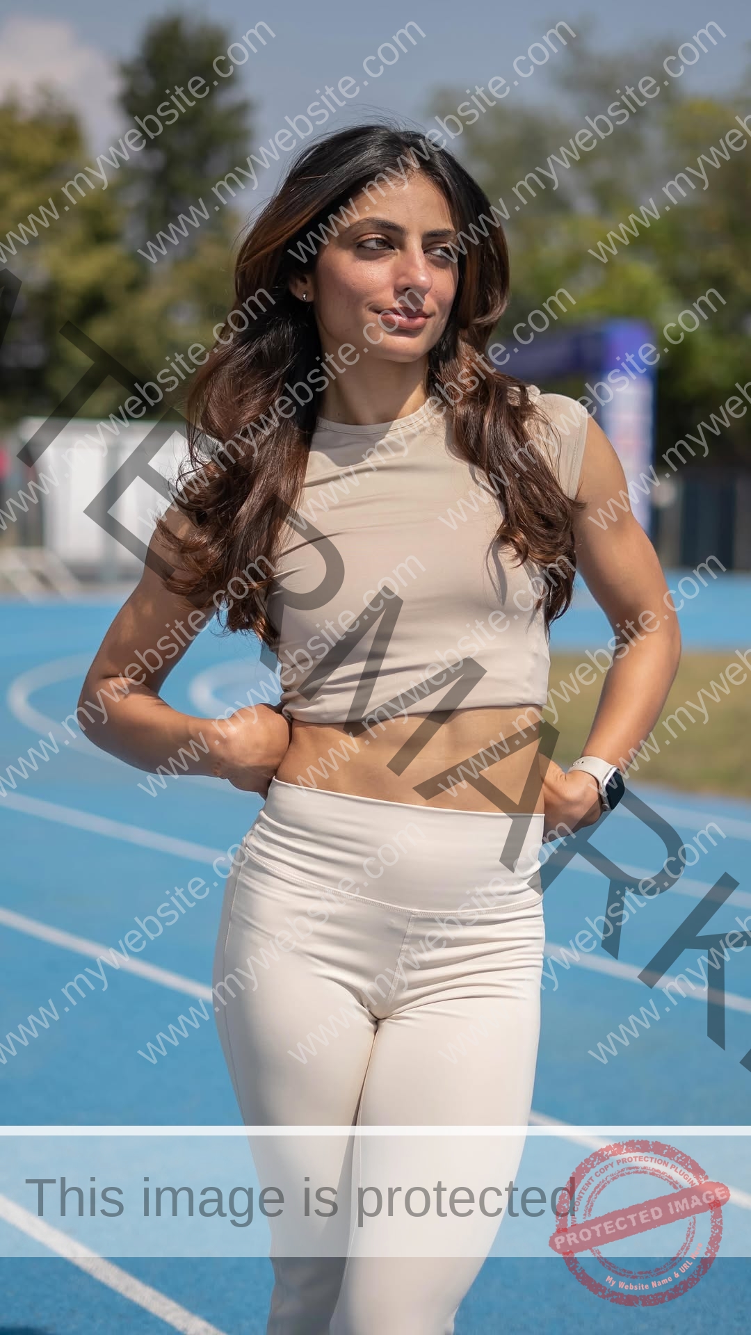 niharika-vashisht-india-niharika.vashhisht-06237 Niharika Vashisht, track and field star from India, walks on a blue track in beige activewear, smiling slightly with trees behind.