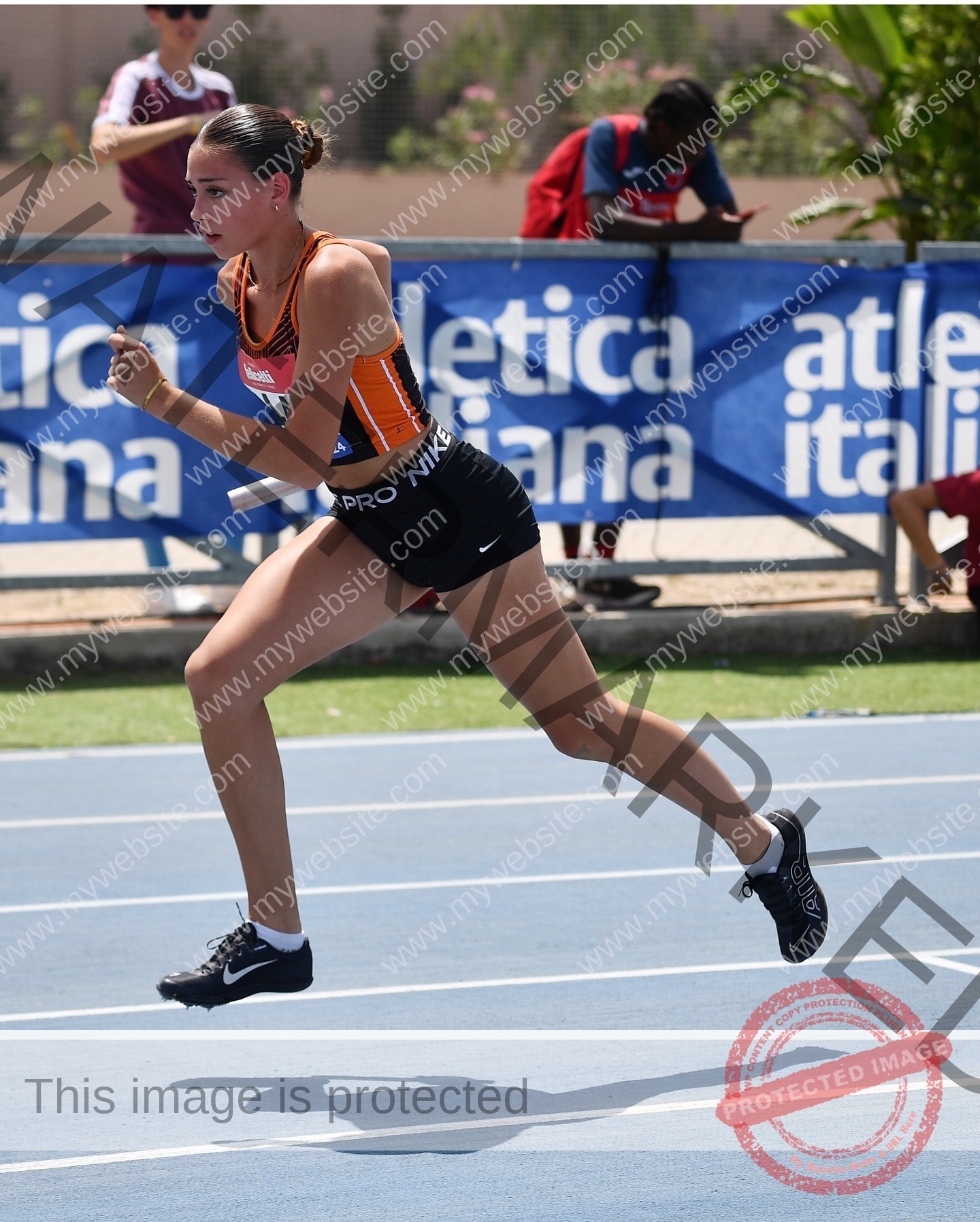 norma-grezio-italy-norma.grezio-u18-xx-07568 Norma Grezio, track and field star from Italy, sprints on a blue track in orange and black, baton in hand; banner and fans behind.