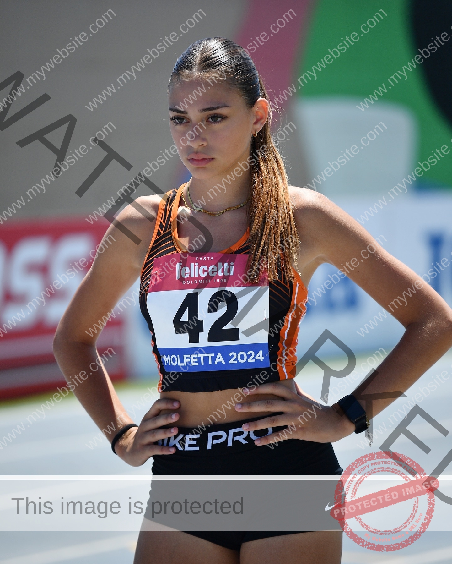norma-grezio-italy-norma.grezio-u18-xx-07569 Norma Grezio, track and field star from Italy, in black and orange gear stands on track, hands on hips, bib 42, Molfetta 2024.