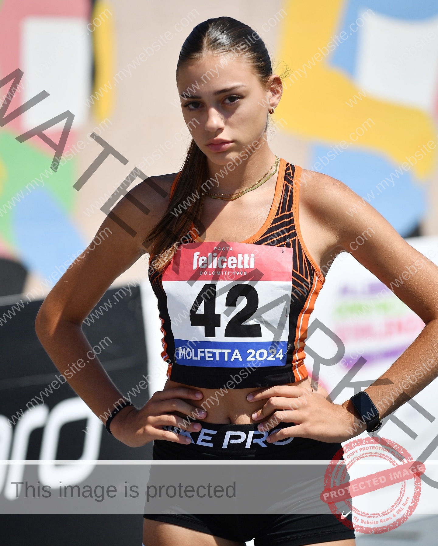 norma-grezio-italy-norma.grezio-u18-xx-07570 Norma Grezio, track and field star from Italy, stands confidently in running gear with number 42 at “Molfetta 2024,” vibrant art behind.