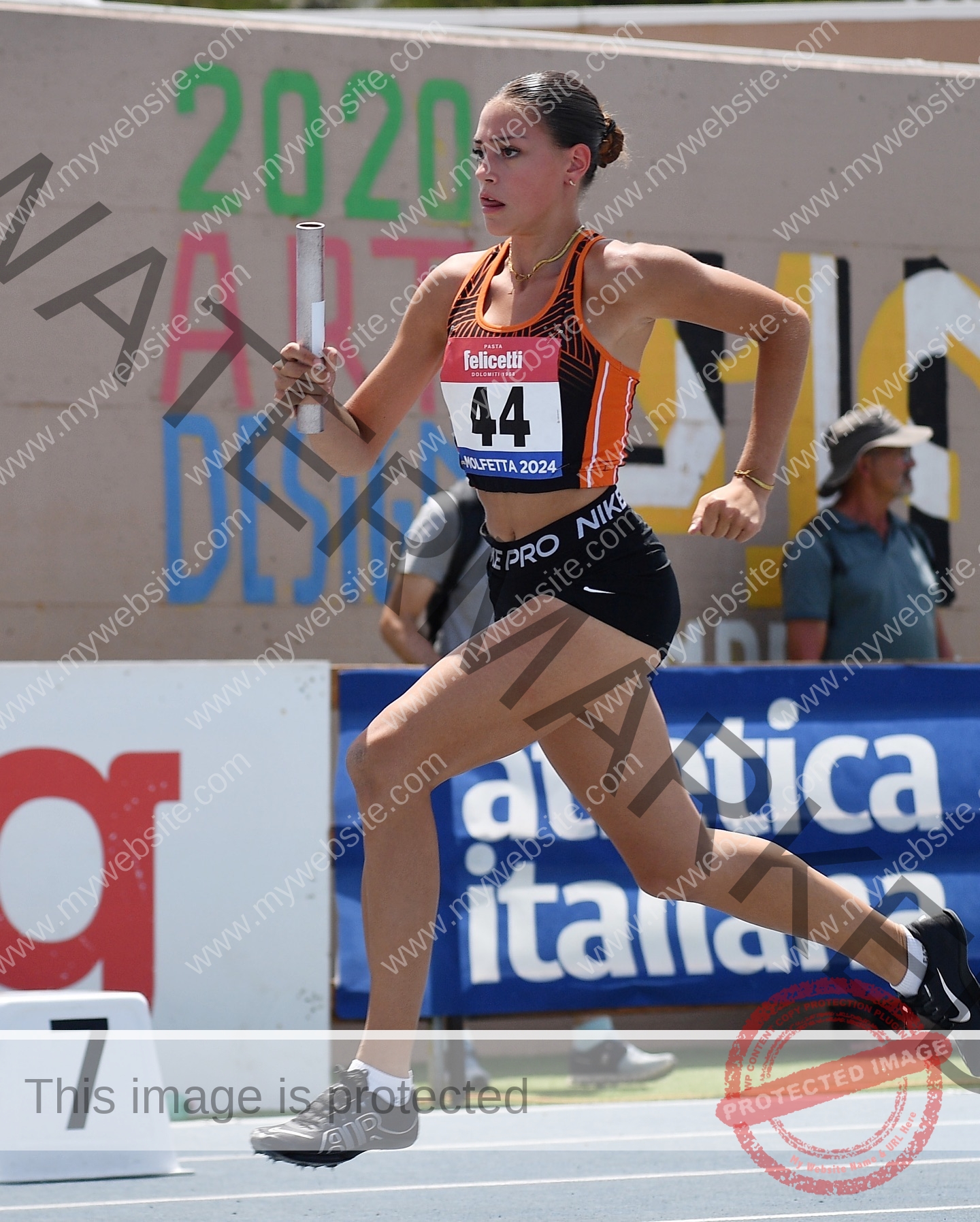 norma-grezio-italy-norma.grezio-u18-xx-07571 Norma Grezio, track and field star from Italy runs mid-stride with baton, in orange/black uniform and #44 bib, looking focused.