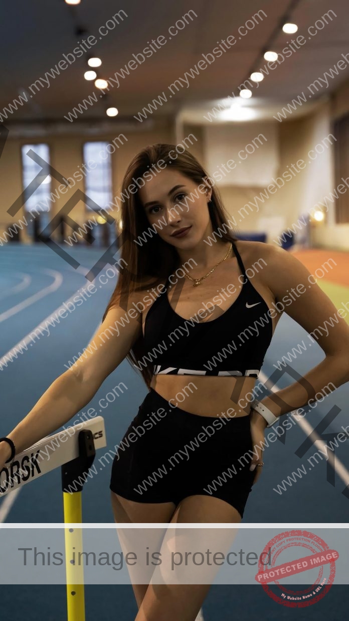 Polina Knoroz, track and field star from Russia, poses confidently indoors by a hurdle on a running track with large windows behind.