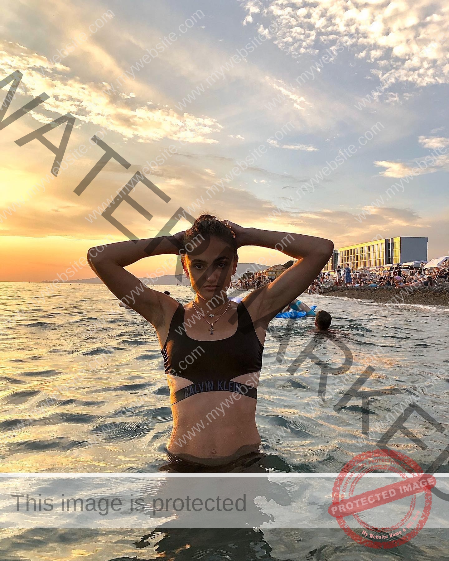 polina-tkalich-russia-p_a_tkalich-10620 Polina Tkalich, track and field star from Russia stands waist-deep in the ocean at sunset, hands on head, black sports bra on.