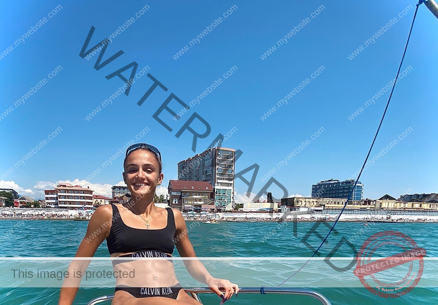 polina-tkalich-russia-p_a_tkalich-10636 Polina Tkalich, track and field star from Russia, in a black Calvin Klein bikini sits on a boat by blue water and seaside buildings.