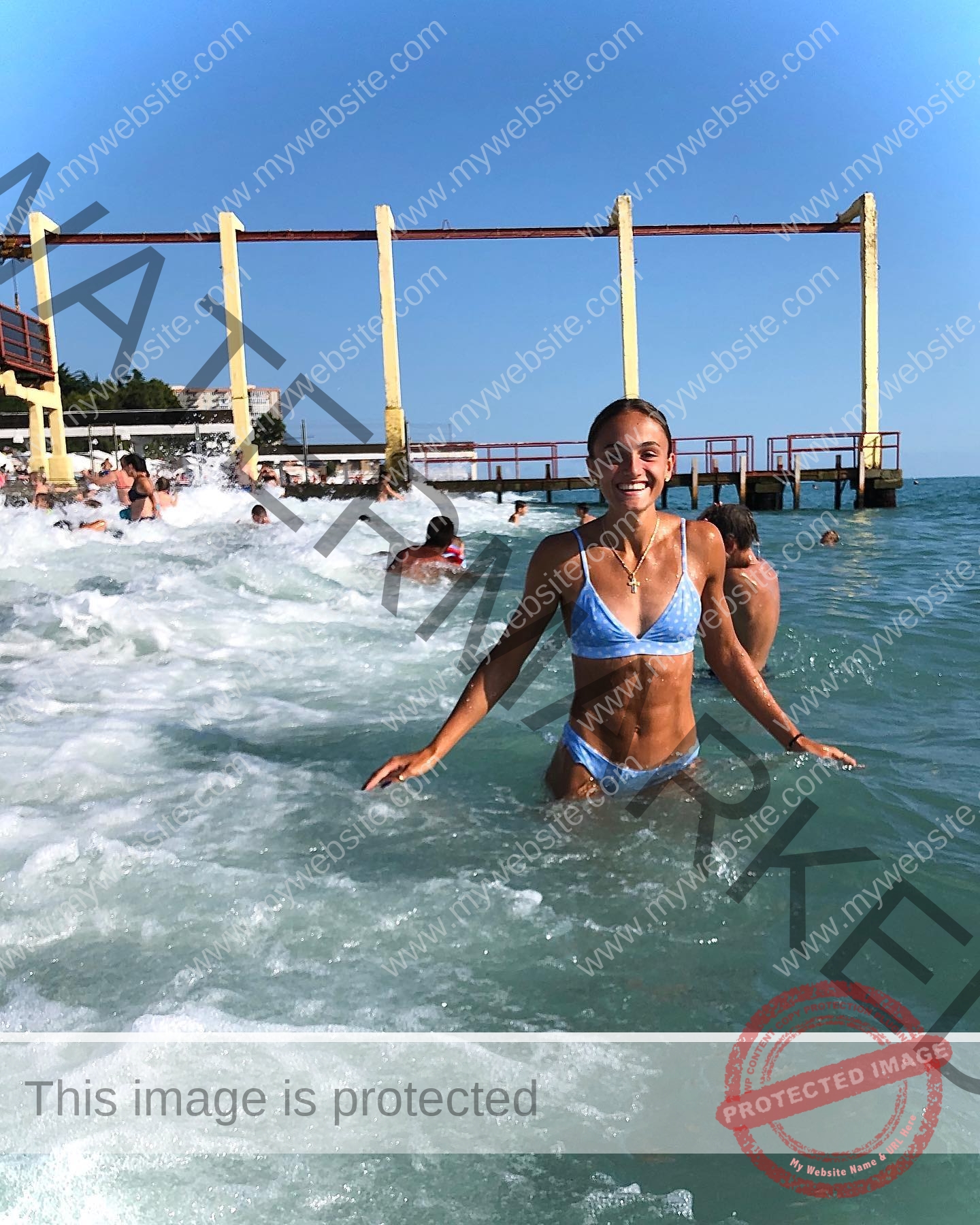 polina-tkalich-russia-p_a_tkalich-10657 Polina Tkalich, track and field star from Russia, stands smiling in a blue bikini waist-deep in ocean waves near yellow piers.