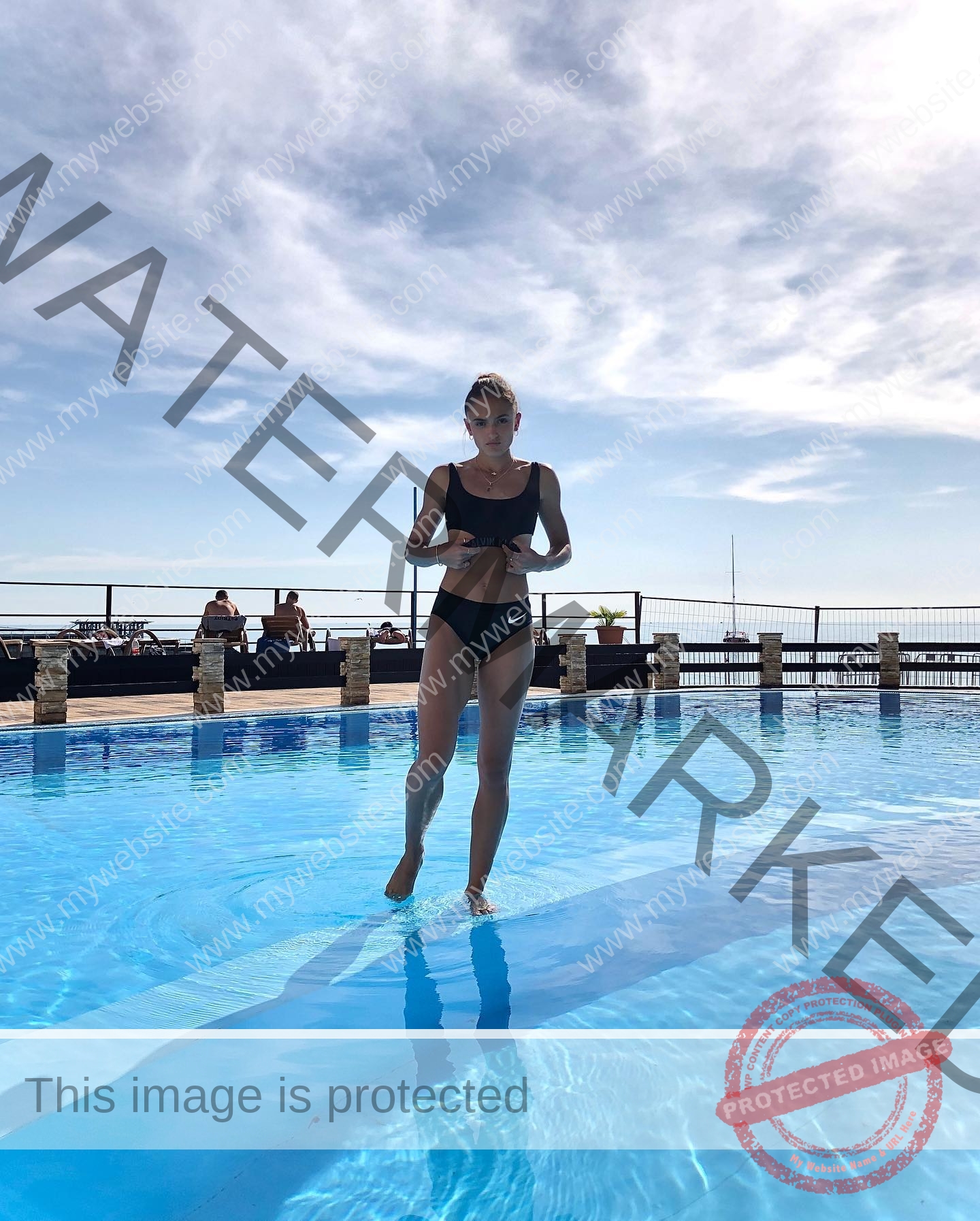 polina-tkalich-russia-p_a_tkalich-10690 Polina Tkalich, track and field star from Russia, walks in a black swimsuit on a blue pool, people by the side, sky behind.