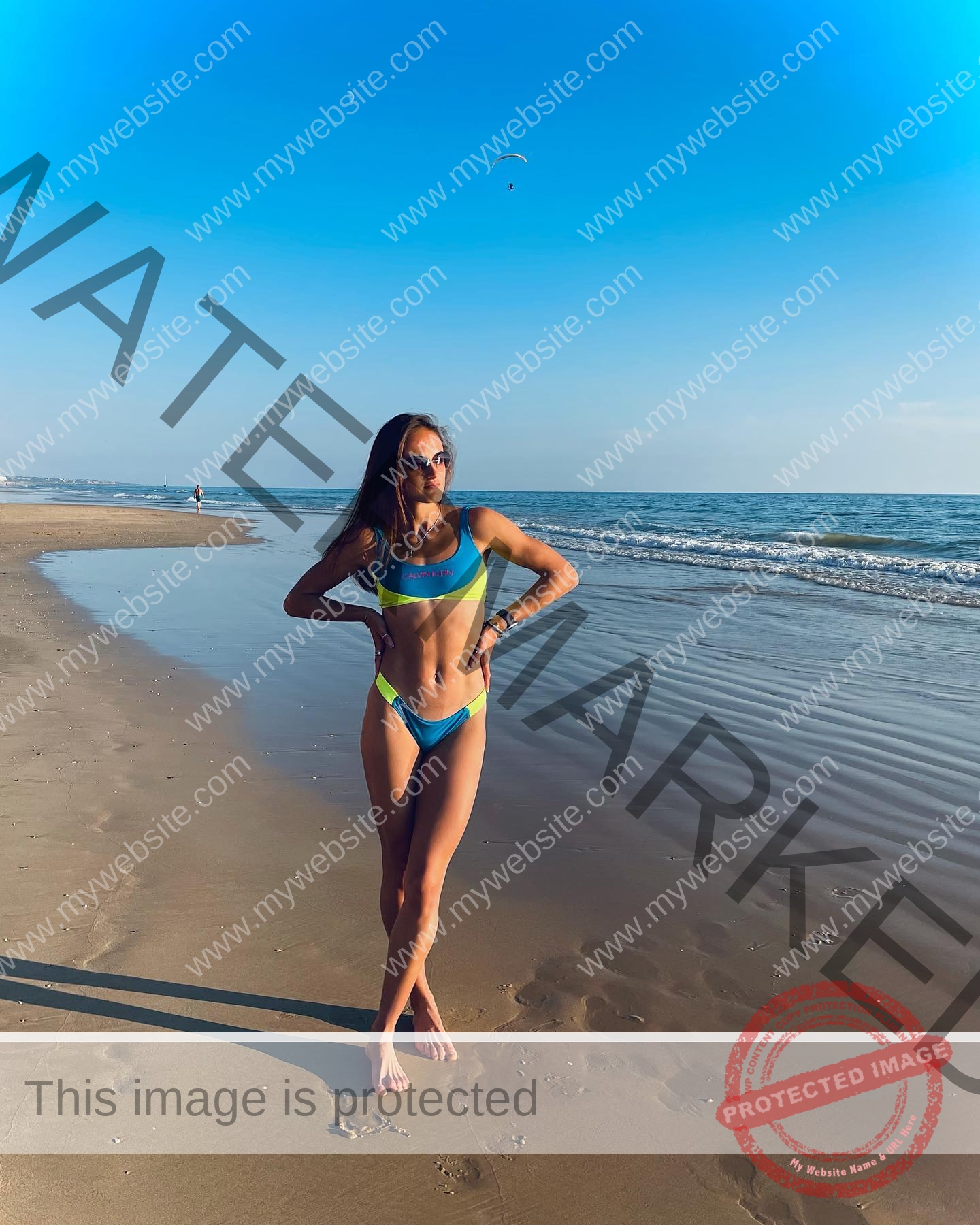 polina-tkalich-russia-p_a_tkalich-10912 Polina Tkalich, track and field star from Russia, poses in a blue-yellow bikini on a sunny beach with sea and distant figure behind.