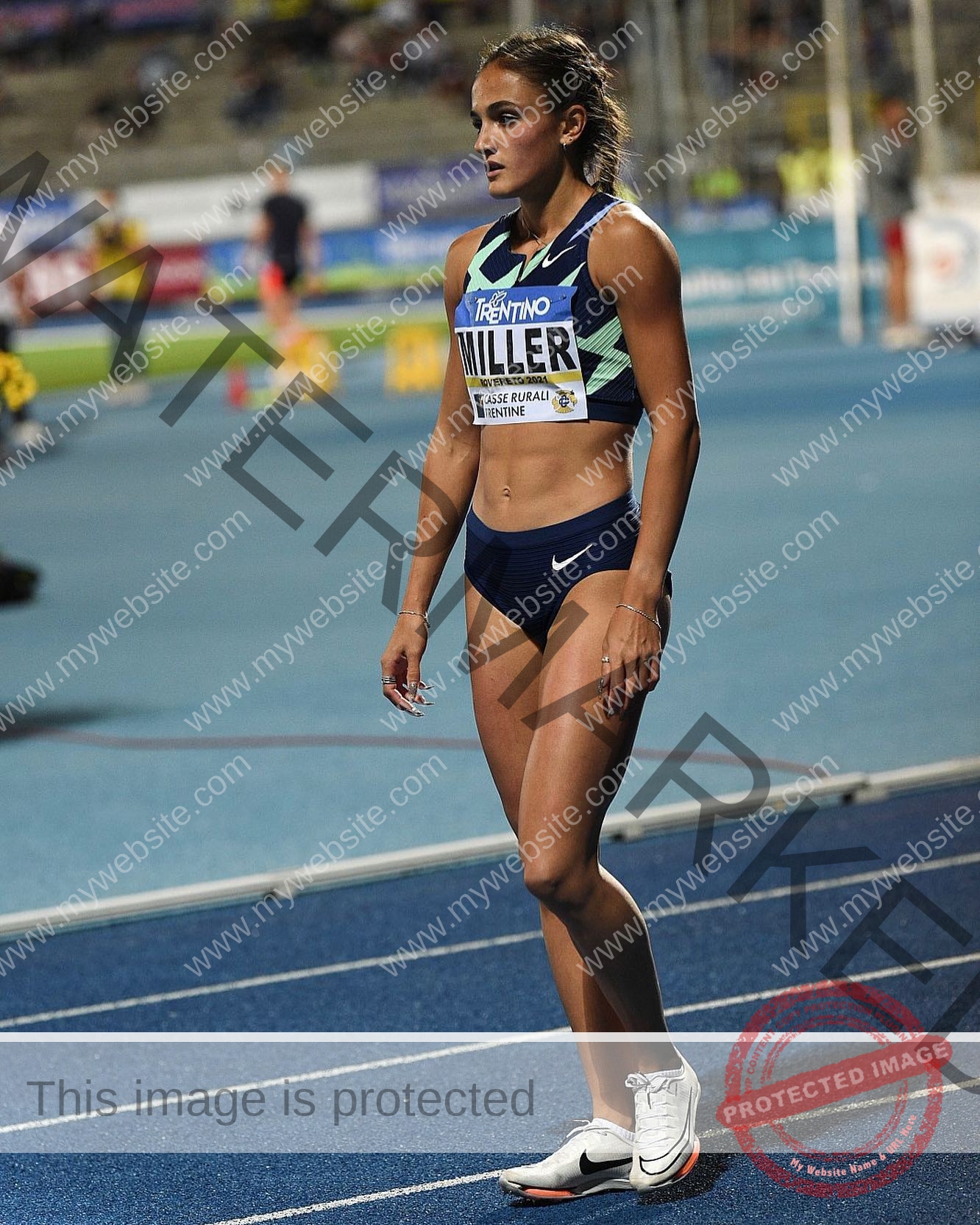 polina-tkalich-russia-p_a_tkalich-10924 Polina Tkalich, track and field star from Russia, in navy sportswear and white shoes stands focused on a blue track under stadium lights.