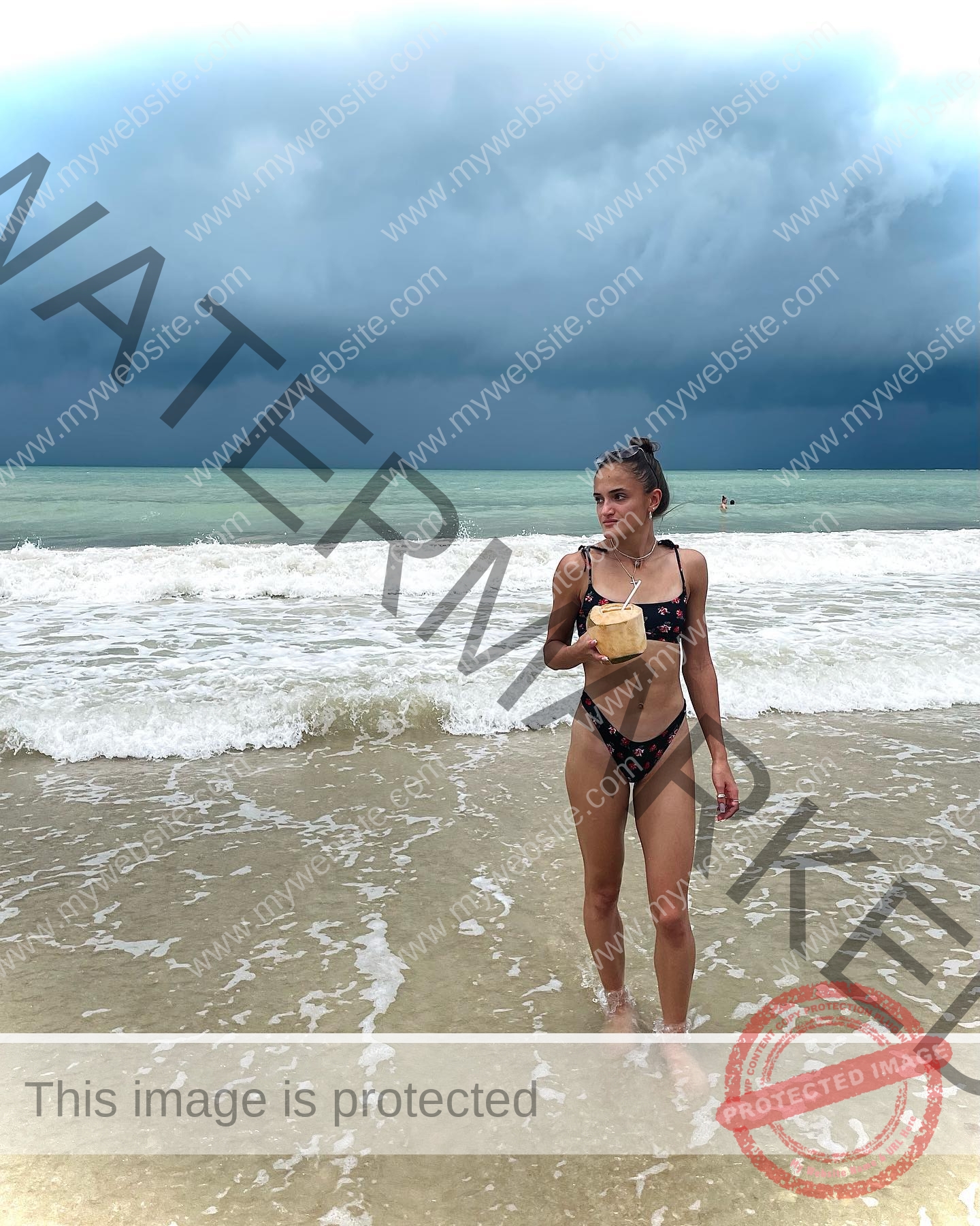 polina-tkalich-russia-p_a_tkalich-11002 Polina Tkalich, track and field star from Russia, stands in shallow ocean water in a black bikini holding a coconut drink; dark waves crash.