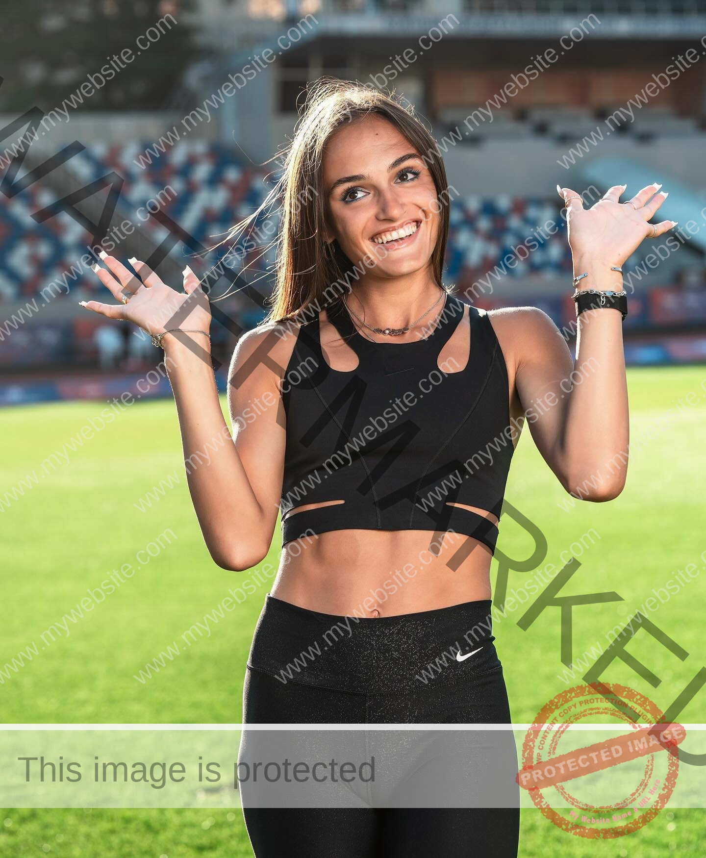 polina-tkalich-russia-p_a_tkalich-11131 Polina Tkalich, track and field star from Russia, smiles in athletic wear, raising her hands playfully on a sports field with stadium seating.