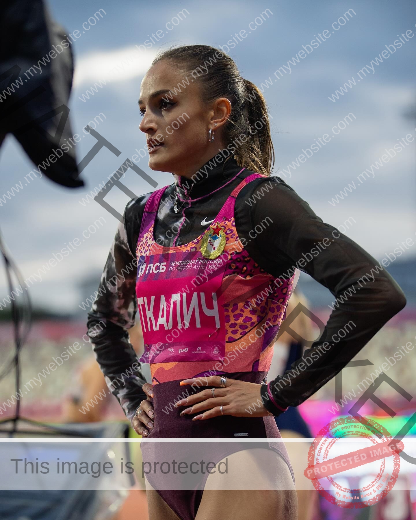 polina-tkalich-russia-p_a_tkalich-11143 Polina Tkalich, track and field star from Russia, stands hands on hips in a colorful uniform at an outdoor event; blurred stadium behind.