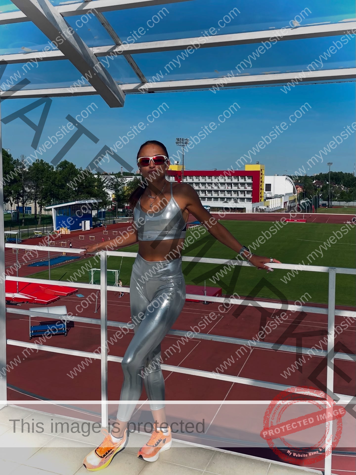 polina-tkalich-russia-p_a_tkalich-11221 Polina Tkalich, track and field star from Russia, in silver athletic gear and orange sneakers poses confidently by a sunny stadium railing.