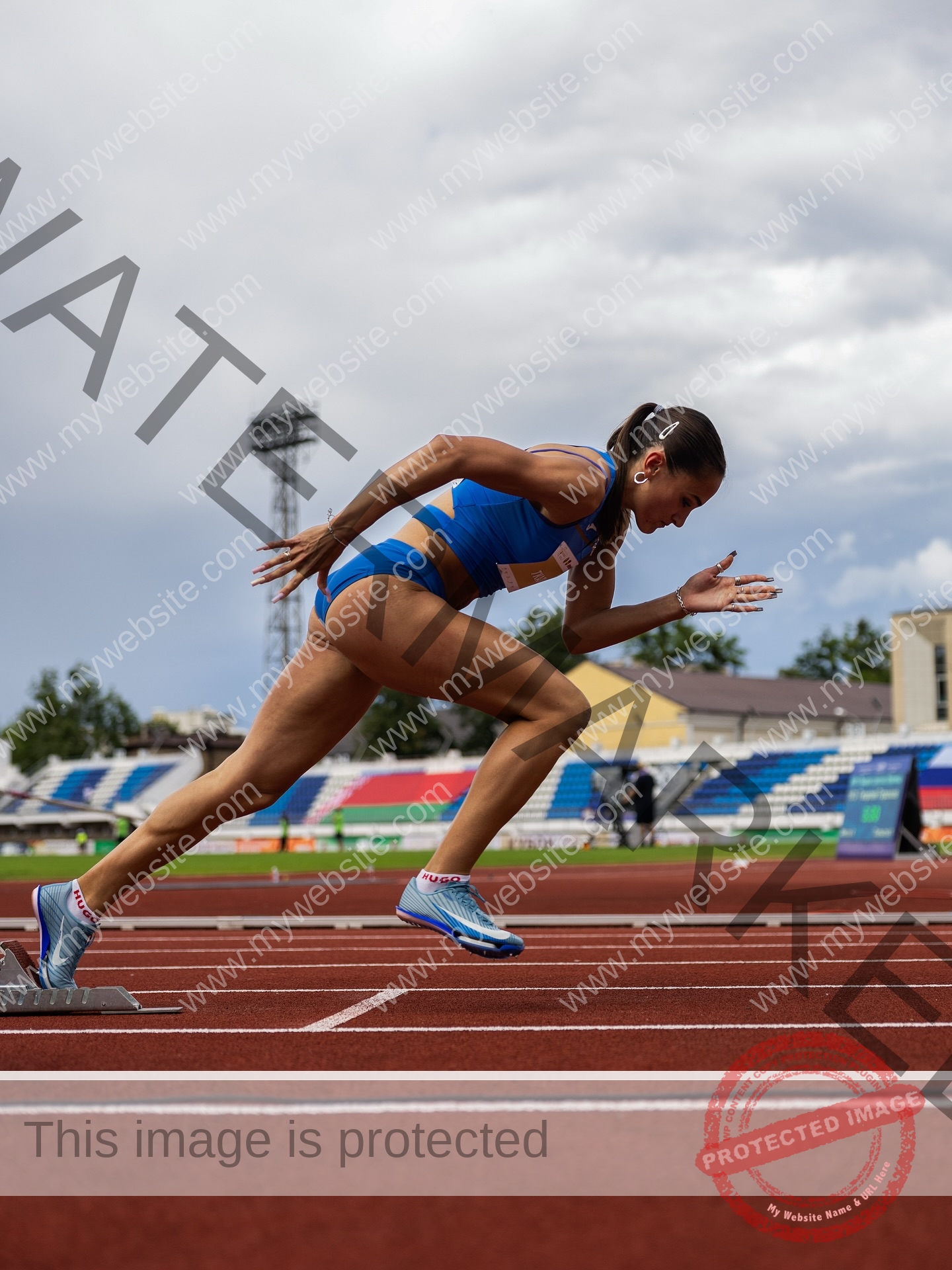 polina-tkalich-russia-p_a_tkalich-11250 Polina Tkalich, track and field star from Russia, in blue sportswear sprints on a red outdoor track, focused under a cloudy sky.