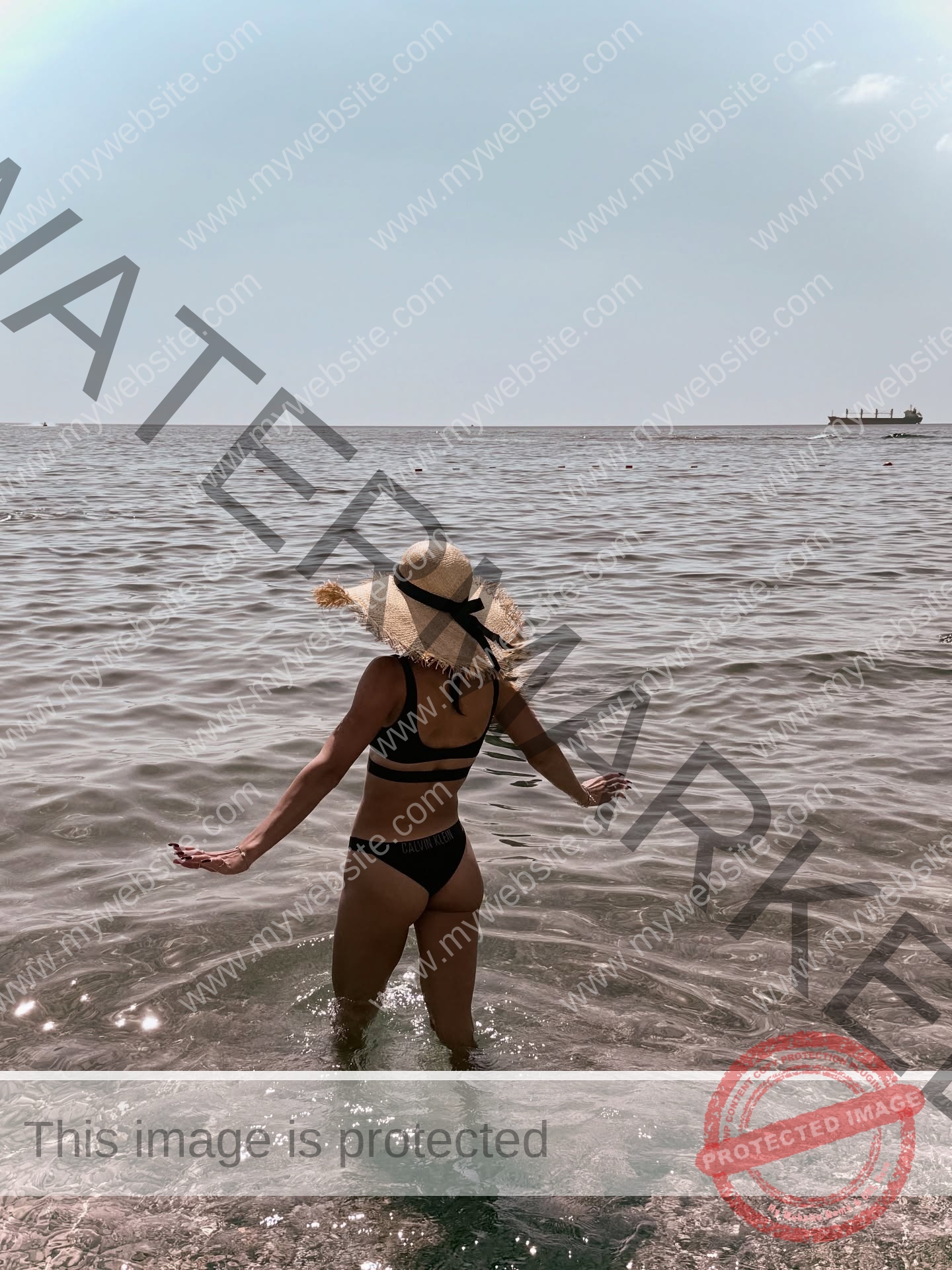polina-tkalich-russia-p_a_tkalich-11299 Polina Tkalich, track and field star from Russia, in a black bikini and hat stands in clear water facing the bright horizon; ship afar.