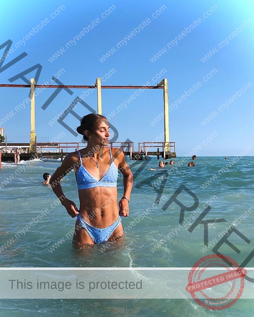 polina-tkalich-russia-p_a_tkalich-11625 Polina Tkalich, track and field star from Russia, in a blue bikini stands in the sea; swimmers and yellow structure behind her.