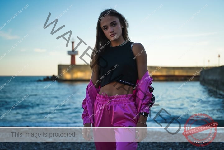 polina-tkalich-russia-p_a_tkalich-11846 Polina Tkalich, track and field star from Russia, in a black crop top and pink outfit stands on a rocky beach at sunset.
