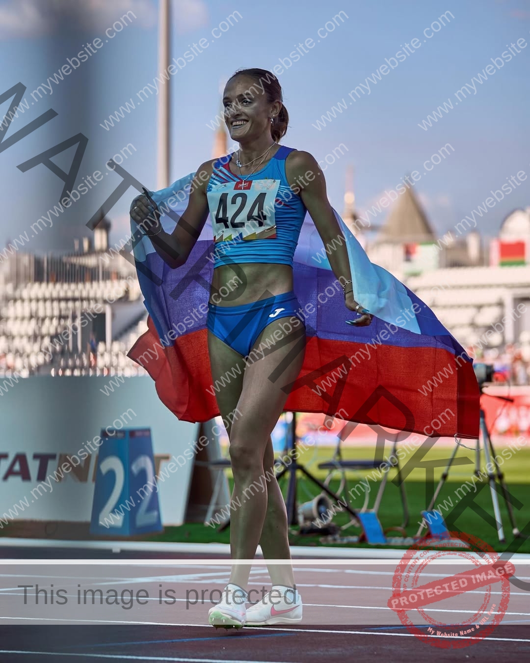 polina-tkalich-russia-p_a_tkalich-12102 Polina Tkalich, track and field star from Russia, smiles in a blue uniform with bib 424, holding a Russian flag on the stadium track.