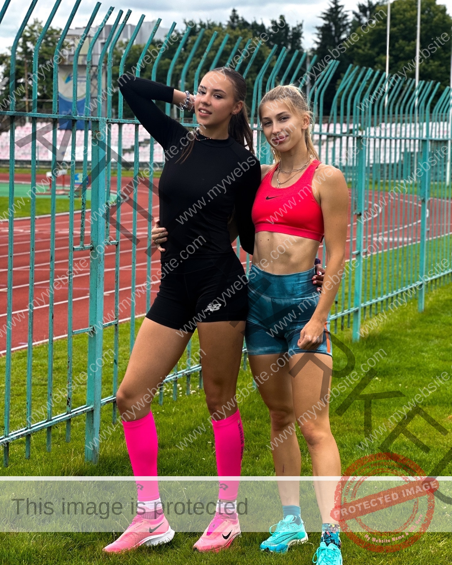 rufina-kotik-ukraine-rufina.kotik14-u18-01864 Rufina Kotik, track and field star from Ukraine, stands on grass with another woman by a track, both smiling near a metal fence.