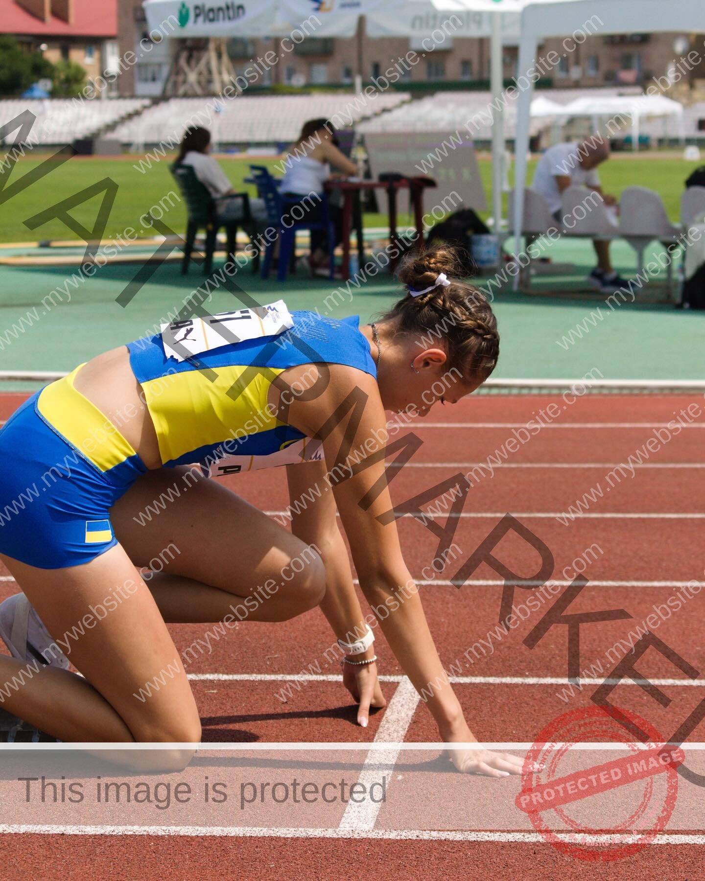 rufina-kotik-ukraine-rufina.kotik14-u18-02016 Rufina Kotik, track and field star from Ukraine, kneels at the starting line in blue and yellow gear; people are in the background.