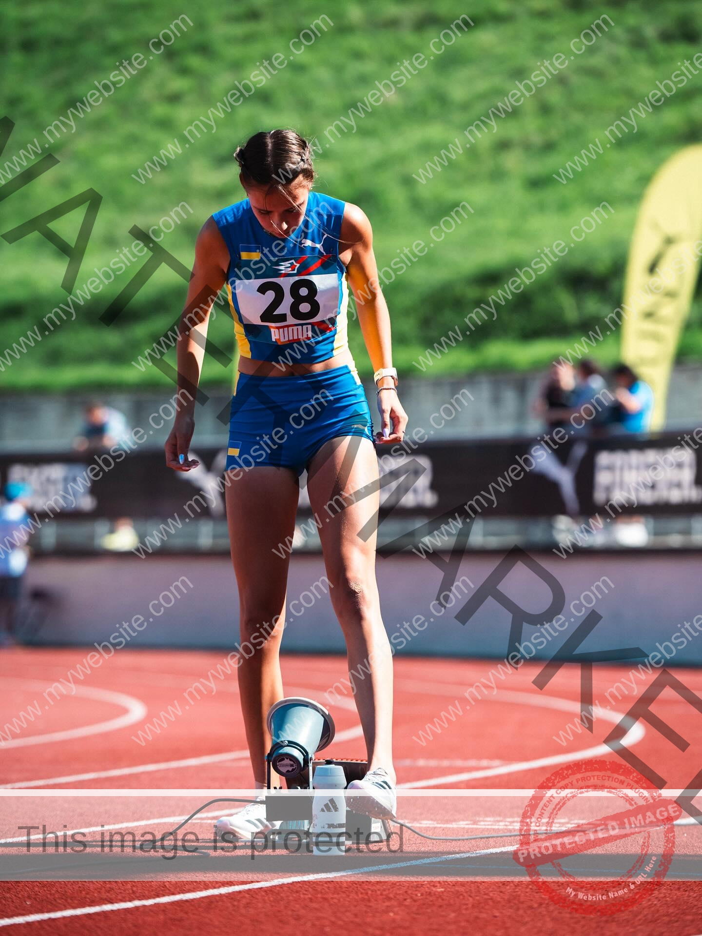 rufina-kotik-ukraine-rufina.kotik14-u18-02049 Rufina Kotik, track and field star from Ukraine, in blue number 28 uniform adjusts starting blocks on a red track with green grass.